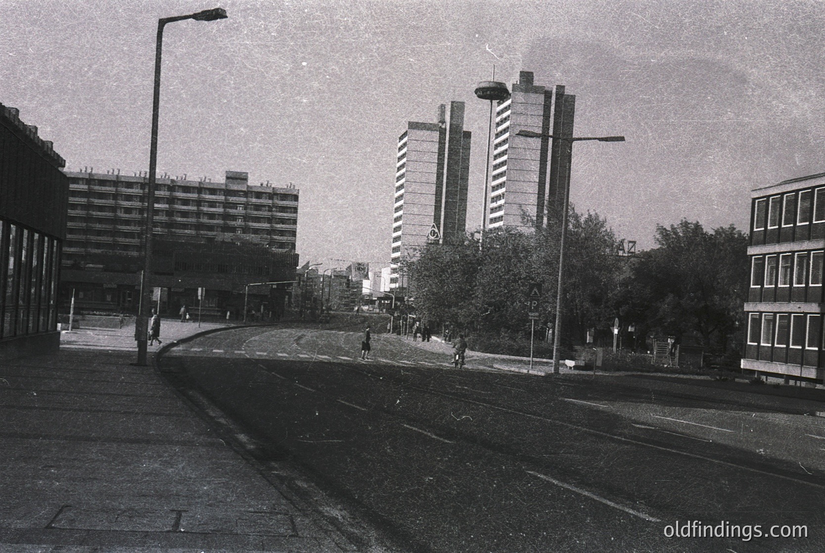 Mid-century urban street scene featuring Brutalist architecture: concrete high-rises with exposed block detailing flank a wide, empty road. Minimal pedestrians and a single streetlamp dominate the scene. Likely Eastern Bloc, 1960s–1970s.