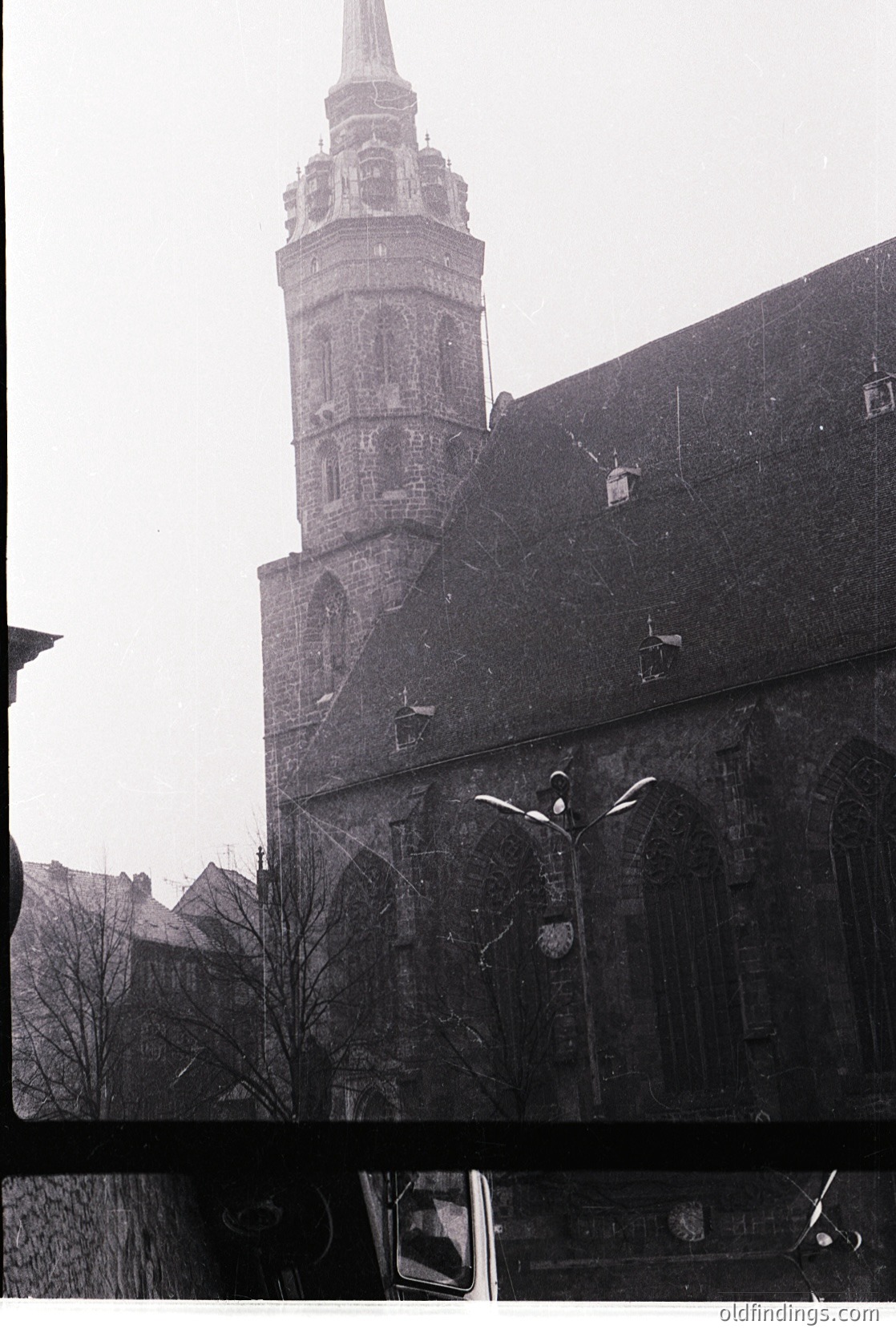 Vintage black-and-white photo of a Gothic-style church tower with ornate brickwork and a pointed spire, flanked by bare trees and street lamps. Partial view of a vintage car in foreground suggests mid-20th century urban setting.