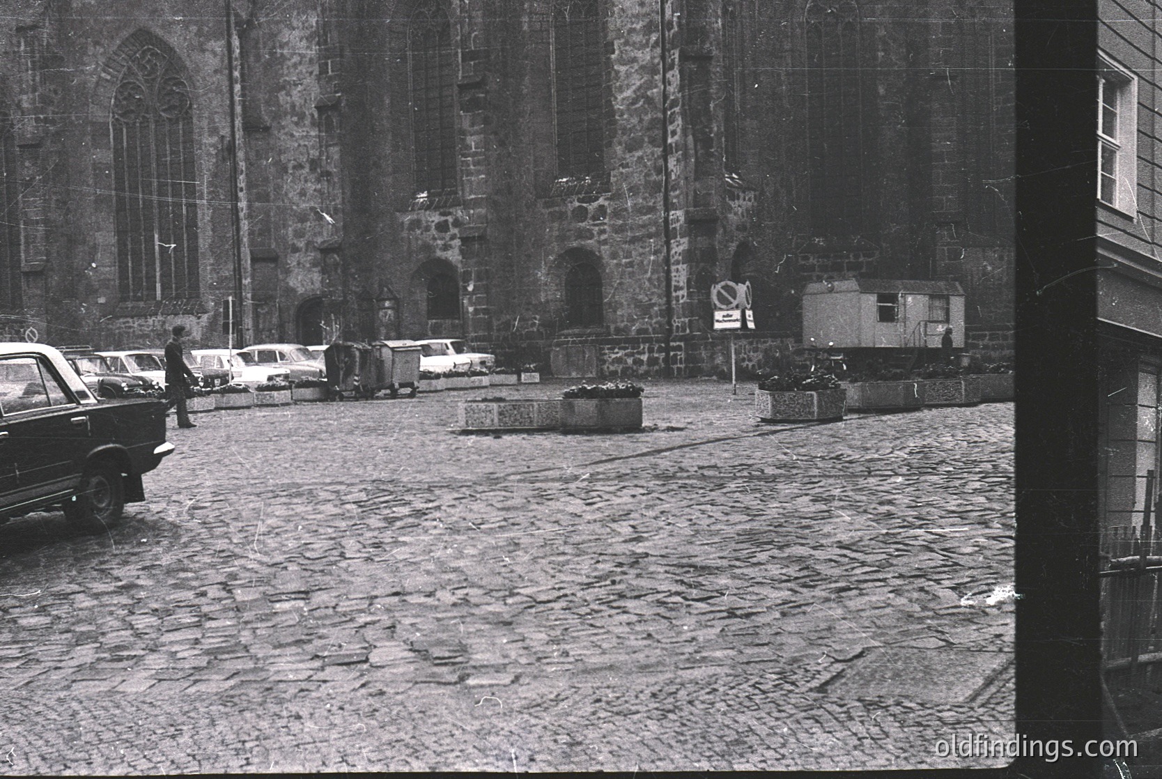 Mid-20th century European plaza with Gothic-style stone church façade featuring pointed arches and weathered brickwork. Cobblestone street with parked vintage cars (1950s–60s era) and a no-parking sign. Temporary barriers and a small portable structure hint at construction or maintenance.