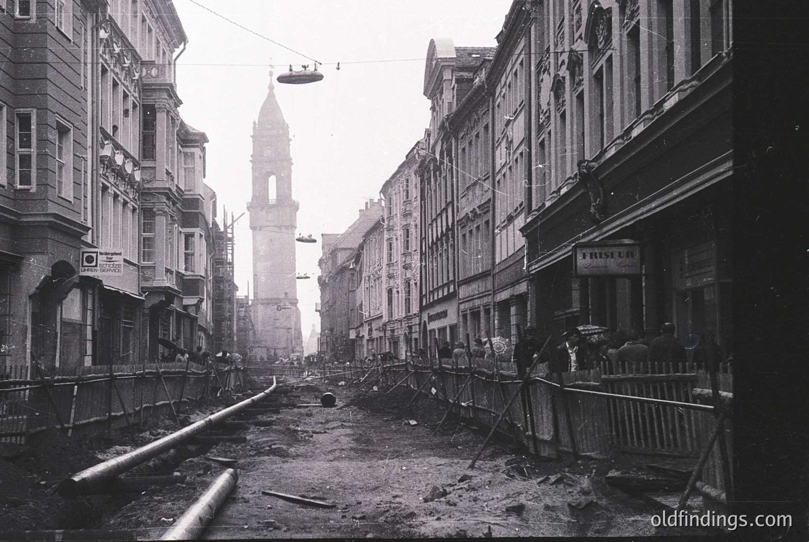 Bomb-damaged street in central European city, likely post-WWII. Ruined buildings flank a debris-strewn thoroughfare, with a suspended cable car ascending toward a partially intact clock tower. Barbed wire and makeshift barriers suggest wartime occupation or reconstruction efforts.