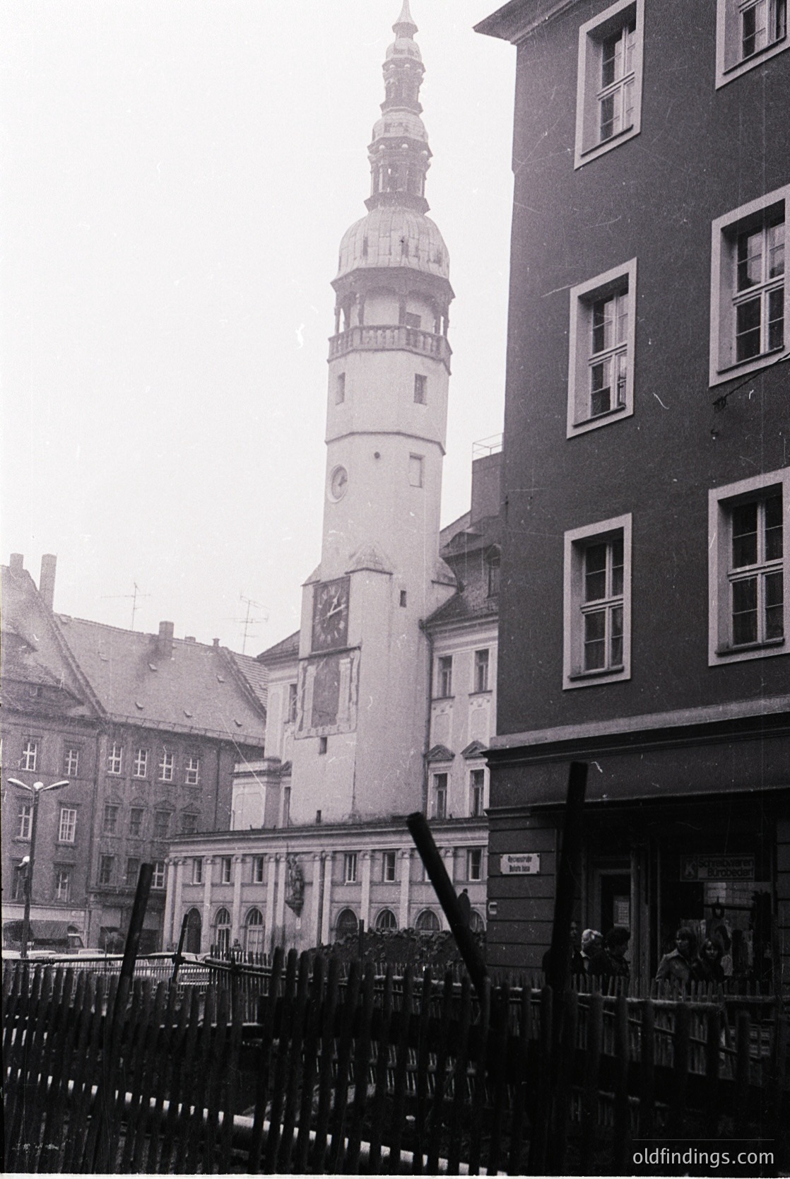 Historic European town square featuring a tall, white Baroque-style church tower with a spire and clock, flanked by multi-story buildings with arched windows. Black wrought-iron fence in foreground. Likely 18th–19th century architecture.