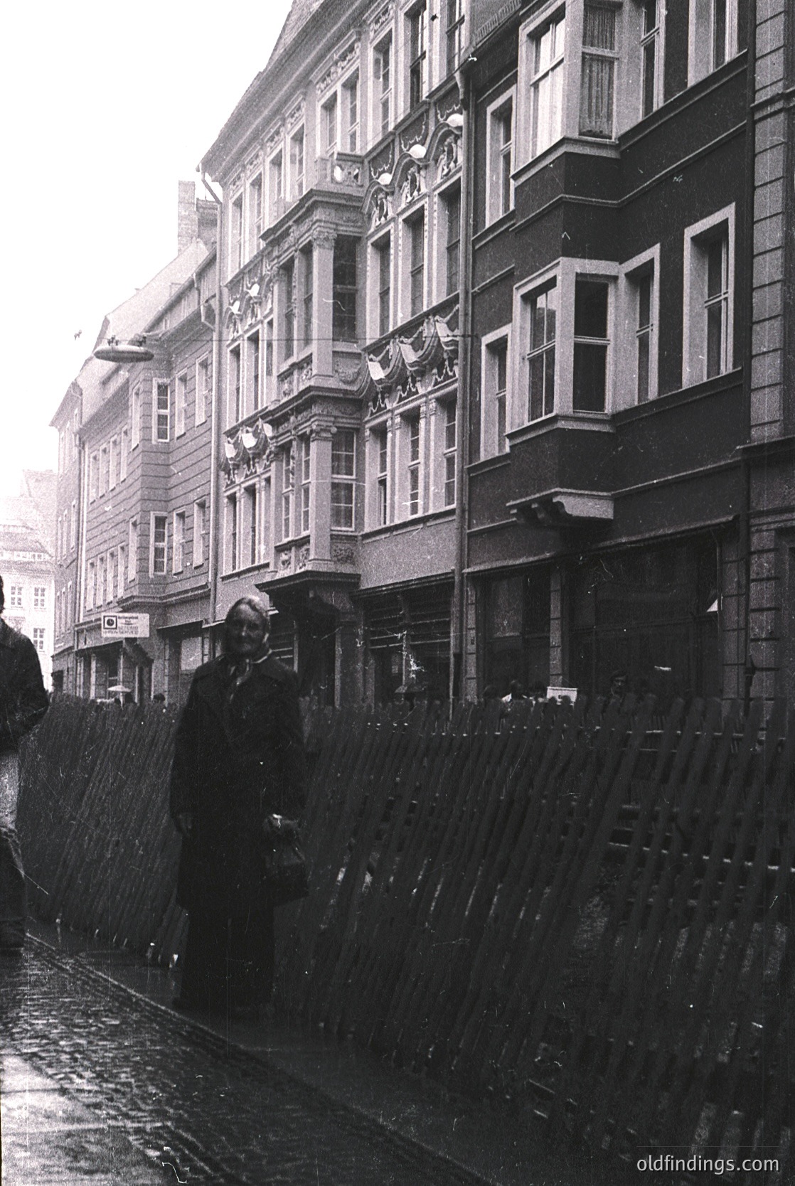 Mid-20th century European street scene featuring classic brick buildings with ornate balconies and large windows. A woman in a long coat and hat stands under a black umbrella, shielding herself from light rain. Wet cobblestones reflect muted light, suggesting a rainy day. Likely