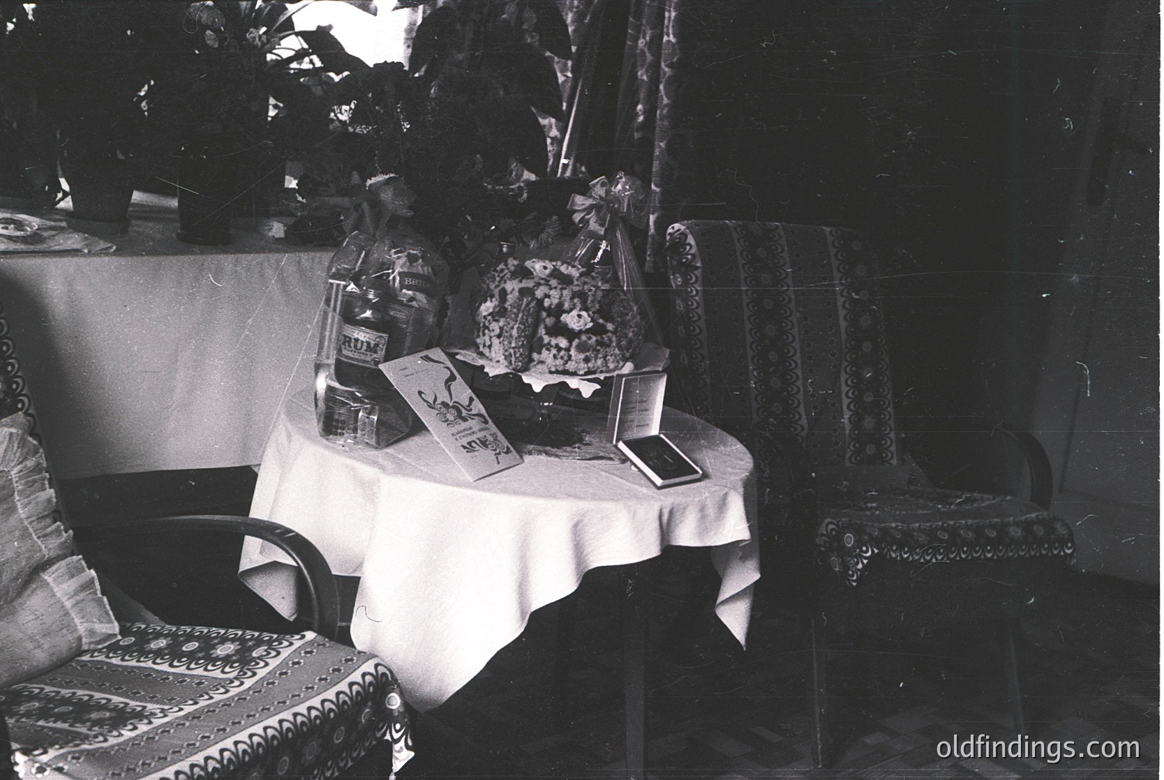 Vintage indoor still-life featuring a small round table draped in a white cloth, adorned with a tiered cake stand holding a layered cake, vintage books, and a framed photograph. Ornate armchairs with intricate embroidery flank the table. Dark, moody lighting enhances textures. Likely mid-20th century domestic setting.