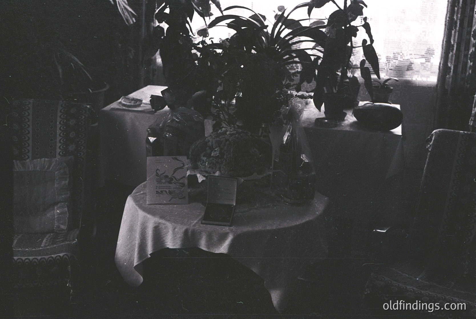 Mid-century dining setup with round table draped in white cloth, surrounded by vintage chairs. Centerpiece features a ceramic vase with greenery, flanked by a framed poster and a bottle. Sunlight streams through a window, illuminating a potted plant and decorative basket. Evokes 1950s–1960s domestic interior design. écor