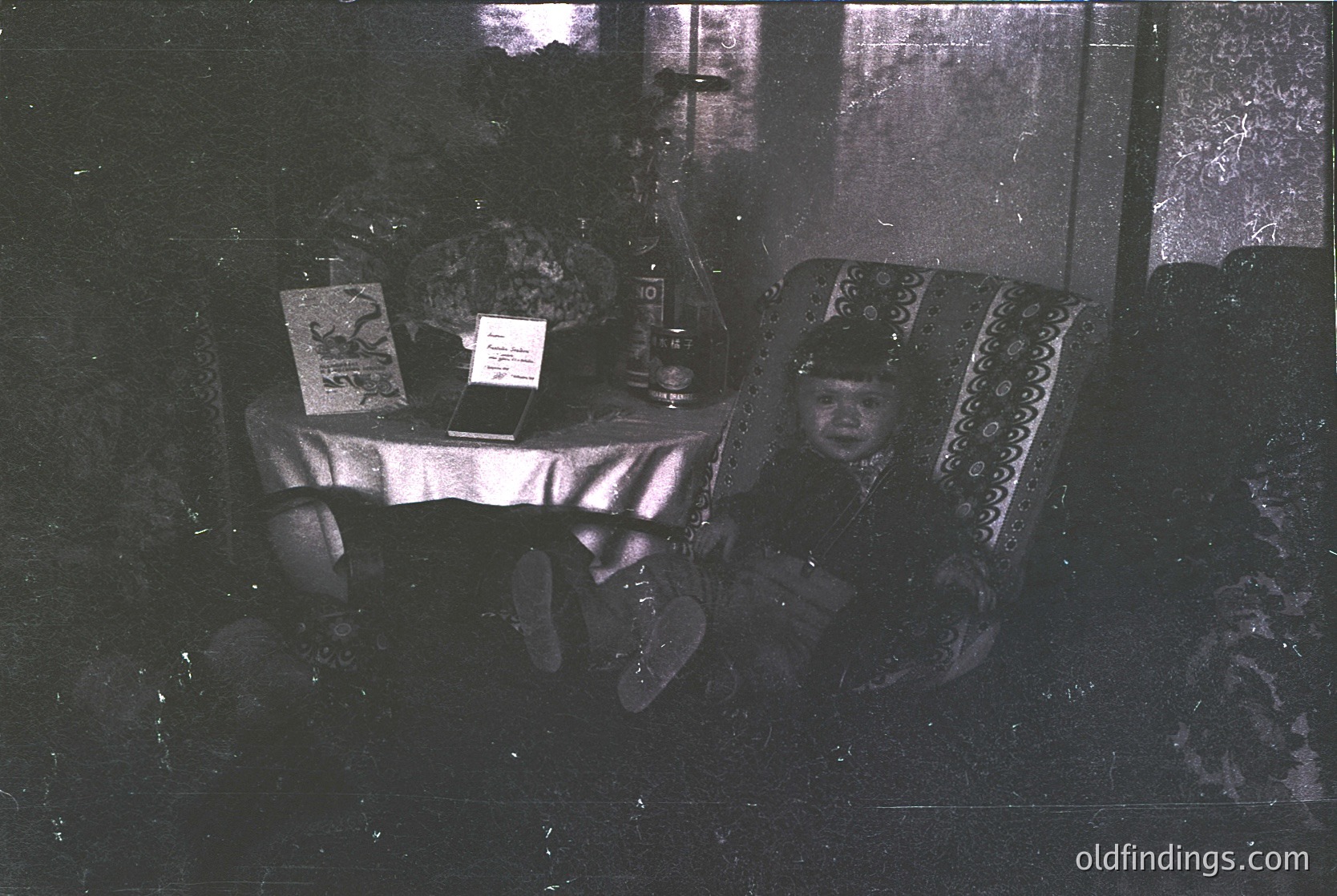 Vintage sepia-toned interior shot of a child seated on ornate, patterned upholstery beside a small table. Table displays a framed poster, a ceramic vase, and a candlestick holder. Dark, textured wall and floor suggest aged, possibly rural or modest home setting. Likely late 19th to early 20th century.