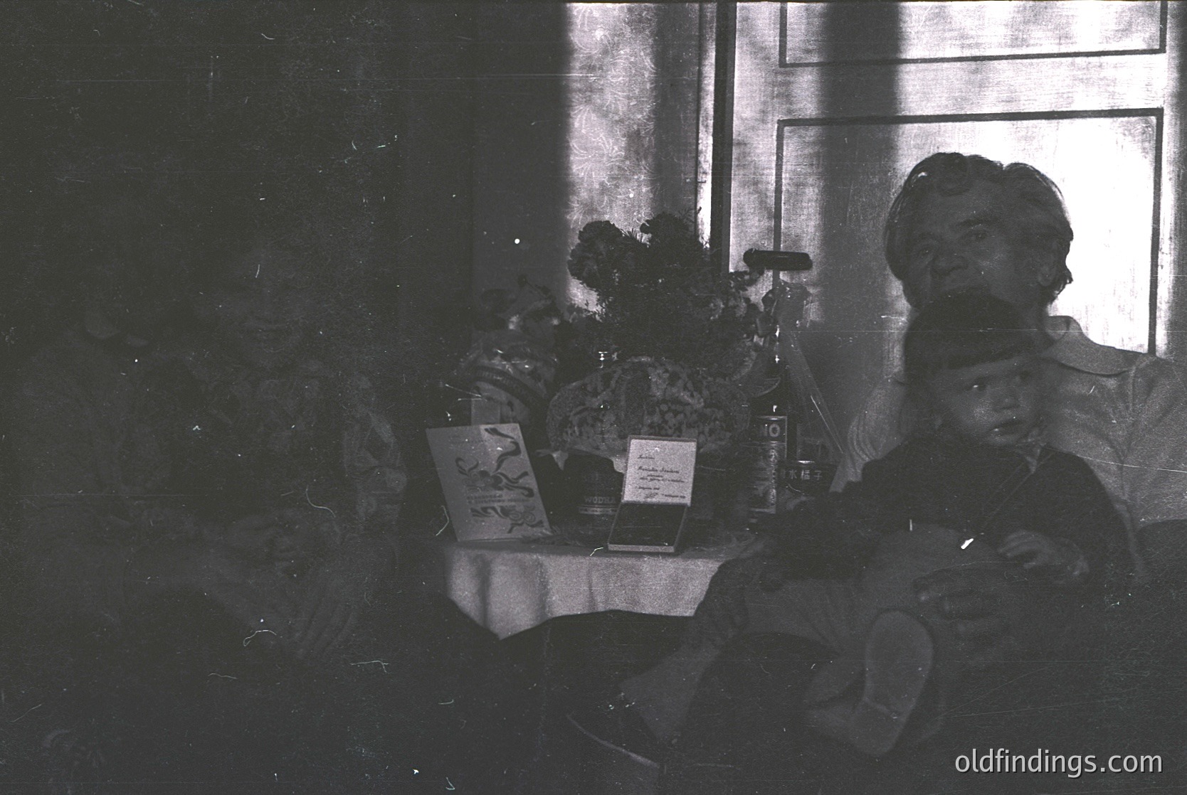 Vintage black-and-white interior shot of a seated man in a military uniform, likely 1940s–1950s. Table beside him holds a book, a small framed photo, and a bottle. Window reveals patterned curtains and a view of a courtyard or street.