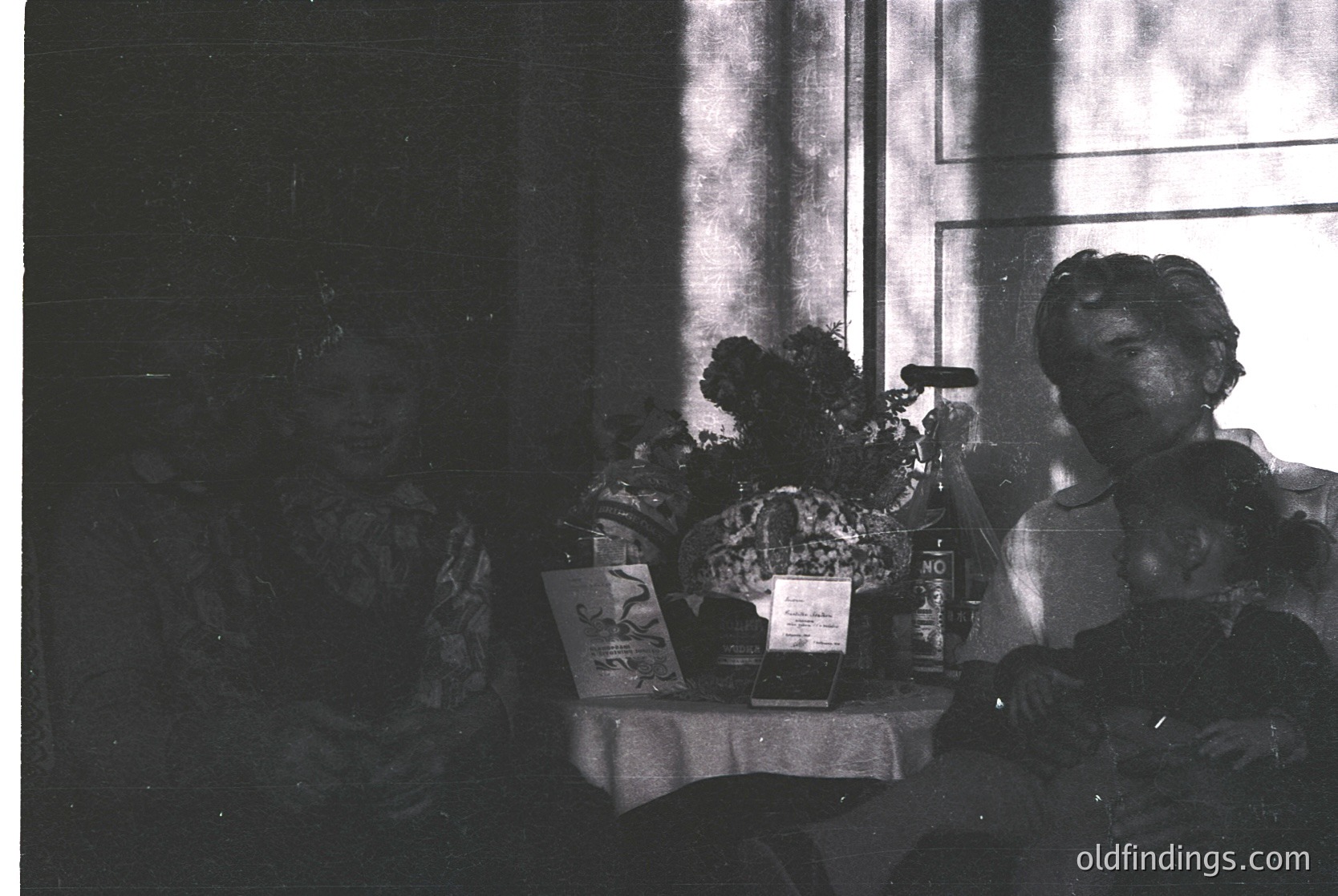 Vintage black-and-white interior shot of a seated woman in a patterned dress, holding a cigarette holder. Center table displays a floral arrangement, vintage books, and a framed photo. Soft natural light filters through a window.