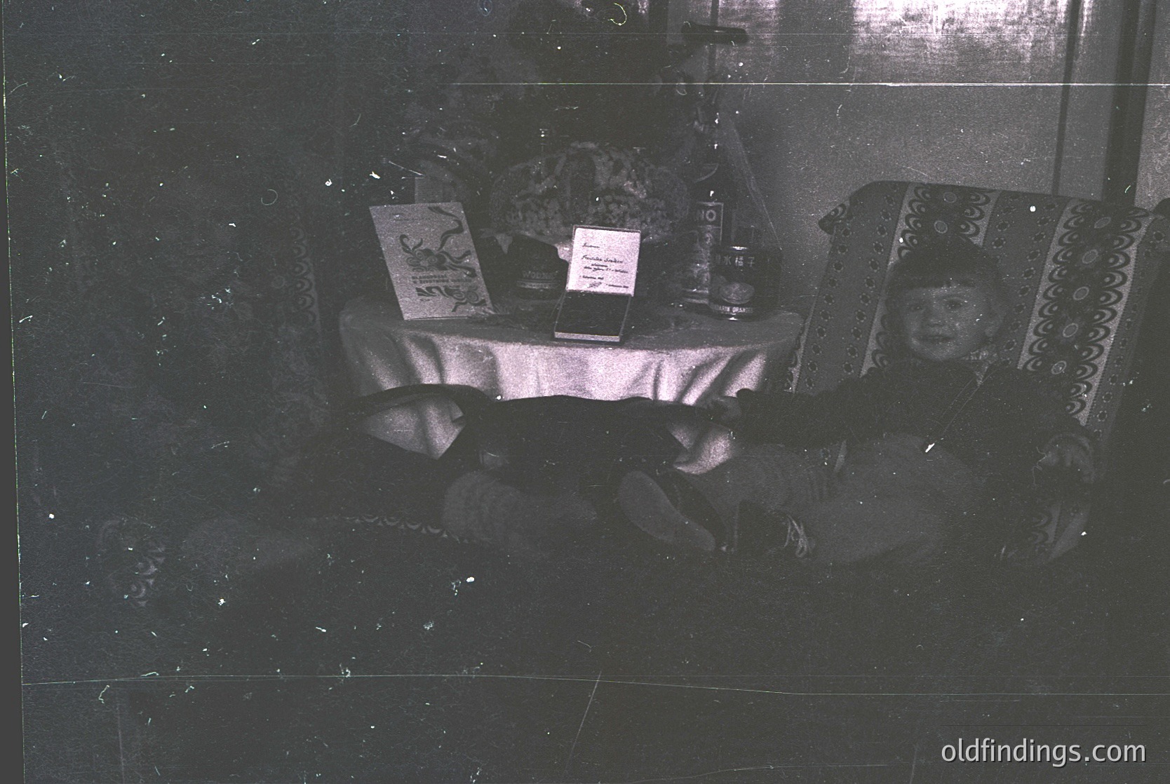 Vintage black-and-white interior shot of a dimly lit room with a low table covered in patterned cloth. Two individuals seated on patterned cushions, one holding a booklet, another a small object. Decorative ceramic vase and framed artwork on table. Reflective surface suggests a mirror or glass barrier. Likely mid-20th century domestic setting.