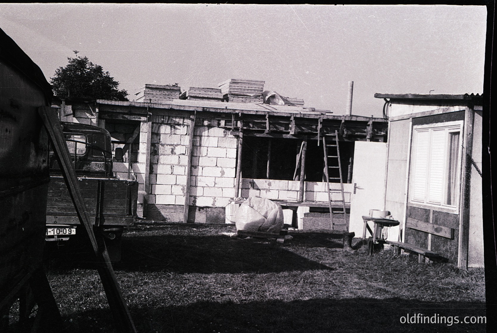 Mid-20th century mobile home under construction, brick exterior with wooden framing visible. Ladder leans against structure; adjacent trailer parked on grass. Simple, utilitarian design reflects post-WWII American housing trends.