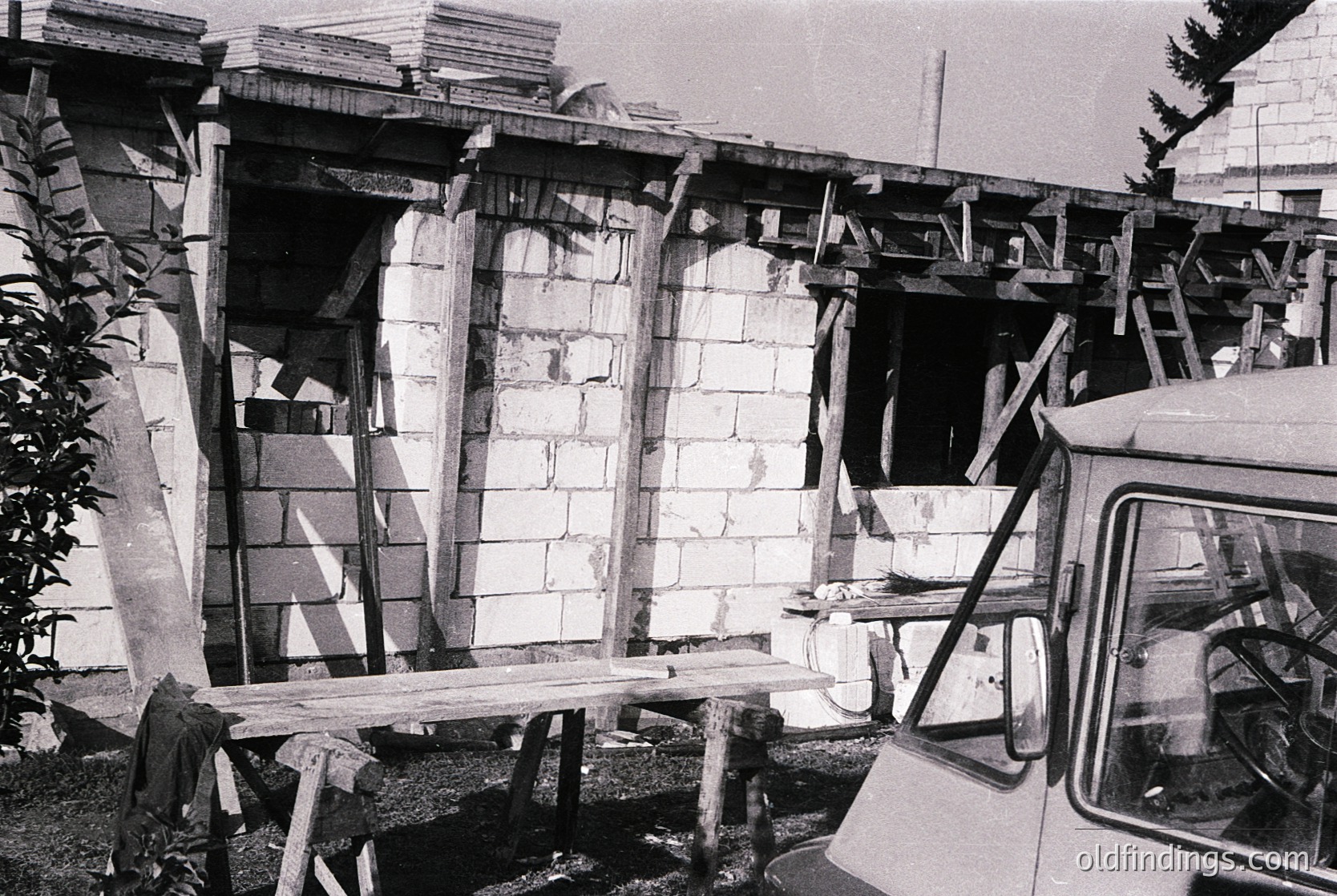 Damaged concrete structure with exposed scaffolding and wooden supports, likely post-earthquake or collapse. Partial view of a vintage truck on right. Urban setting with visible modern buildings in background. Black-and-white, mid-20th century.
