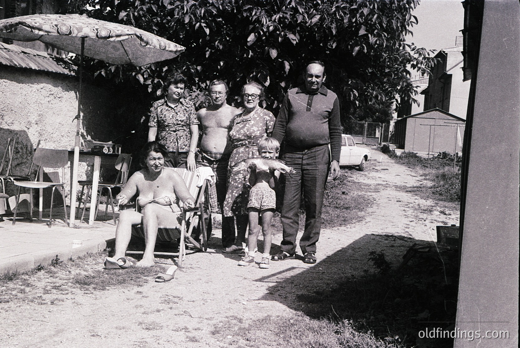 Family portrait from the 1960s–70s, likely Eastern Europe. Five adults and a child pose outdoors near a shaded umbrella and vintage car. Casual beachwear and relaxed postures suggest a summer gathering.