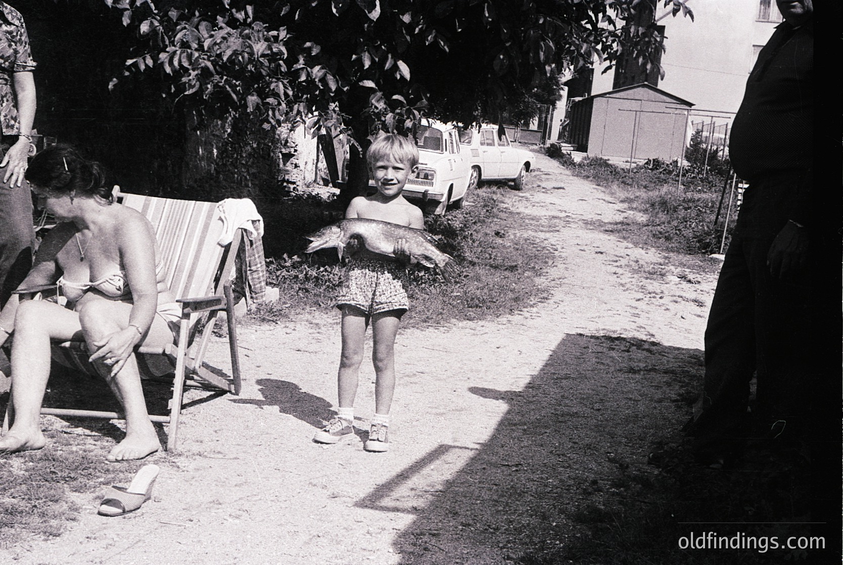 Vintage black-and-white snapshot of a mid-20th-century backyard scene. A young girl in floral shorts and sandals stands holding a stuffed bird, smiling at the camera. A woman in a bikini sits on a folding chair beside her. Classic 1950s-60s cars parked in the background. Residential suburban setting.