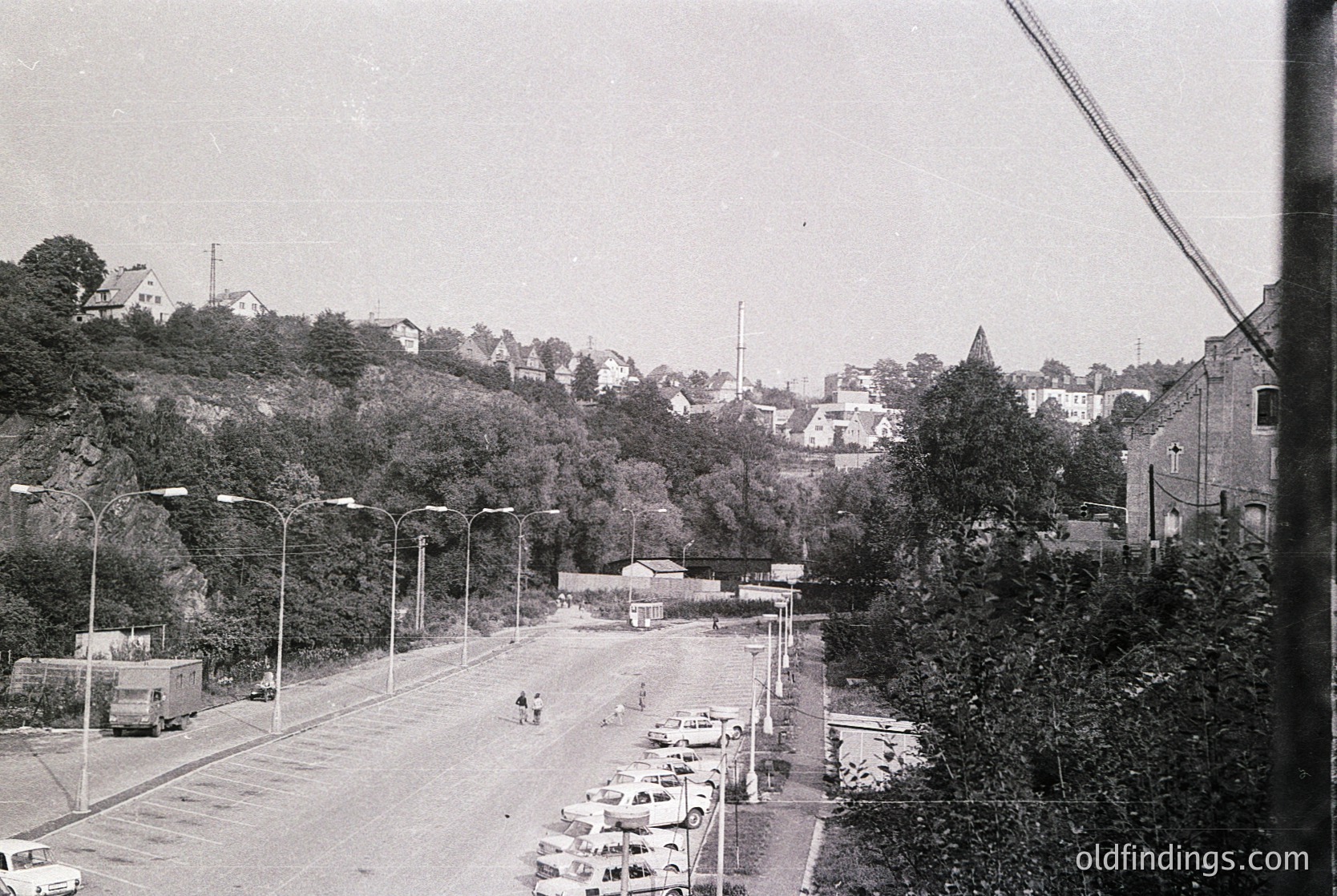 Vintage black-and-white street scene with parked vintage cars, likely 1960s–1970s. Urban landscape featuring a mix of residential buildings, dense greenery, and a roadside market stall. Overcast sky and minimal pedestrians suggest a quiet moment in a European town.