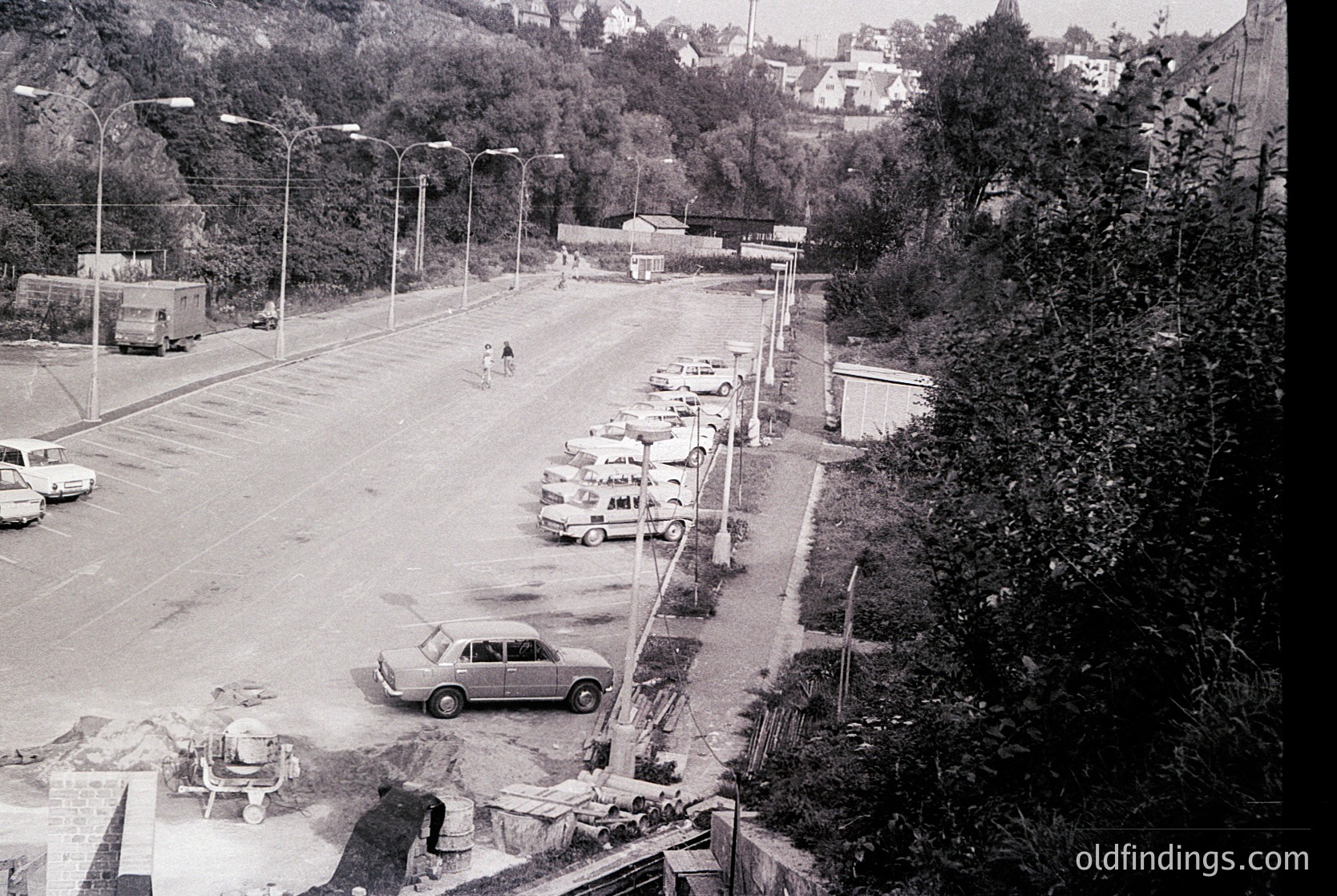Aerial view of a mid-20th-century seaside parking lot with parked cars lining both sides. Concrete barriers and utility poles divide lanes. A lone figure walks near the center, while a parked car drives toward the camera. Dense greenery and residential buildings rise in the background.