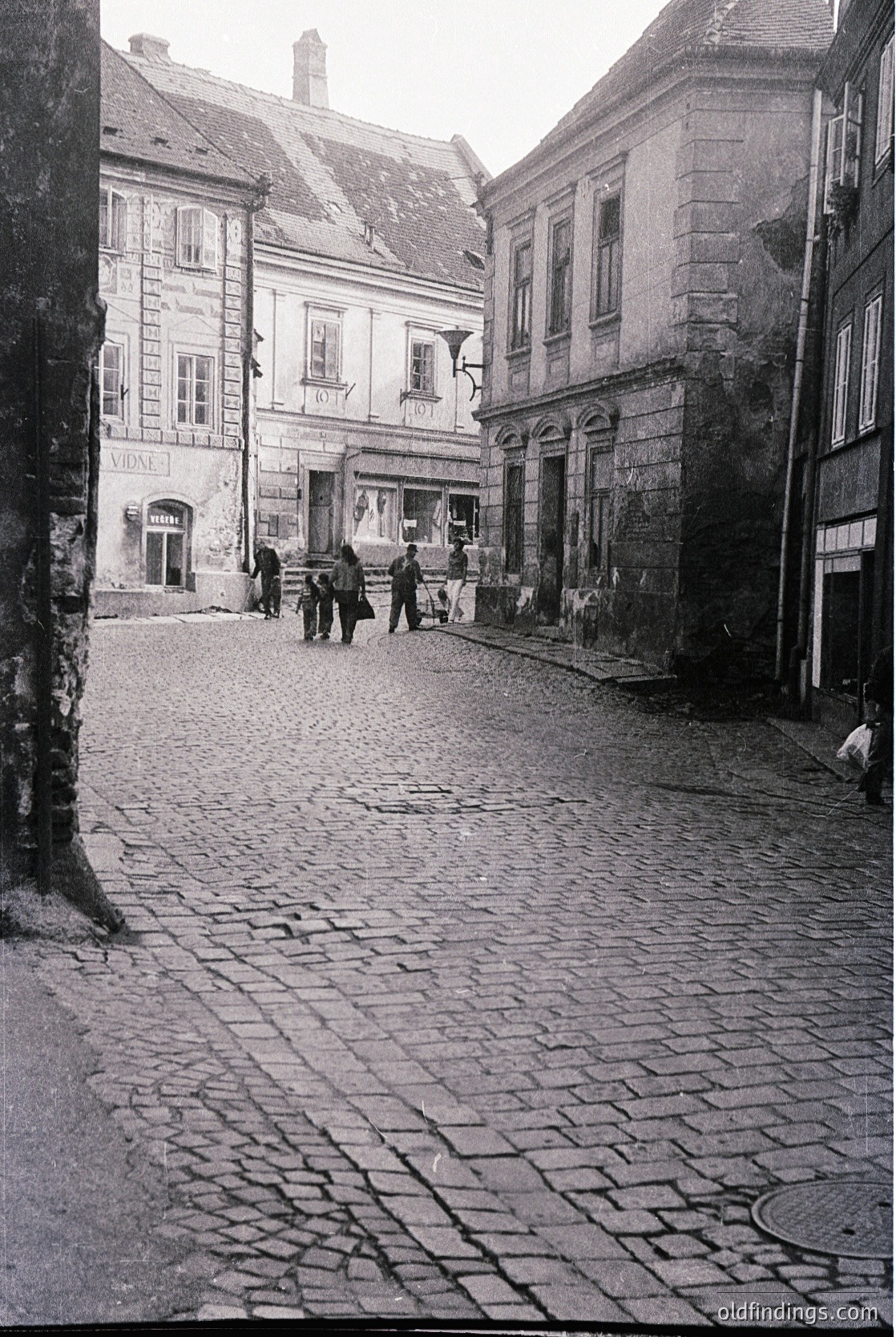 Cobblestone street in a European town with mid-20th century architecture—stone buildings with arched doorways and shuttered windows. Signage in Cyrillic suggests Eastern European locale. People dressed in 1960s-70s attire walking casually.
