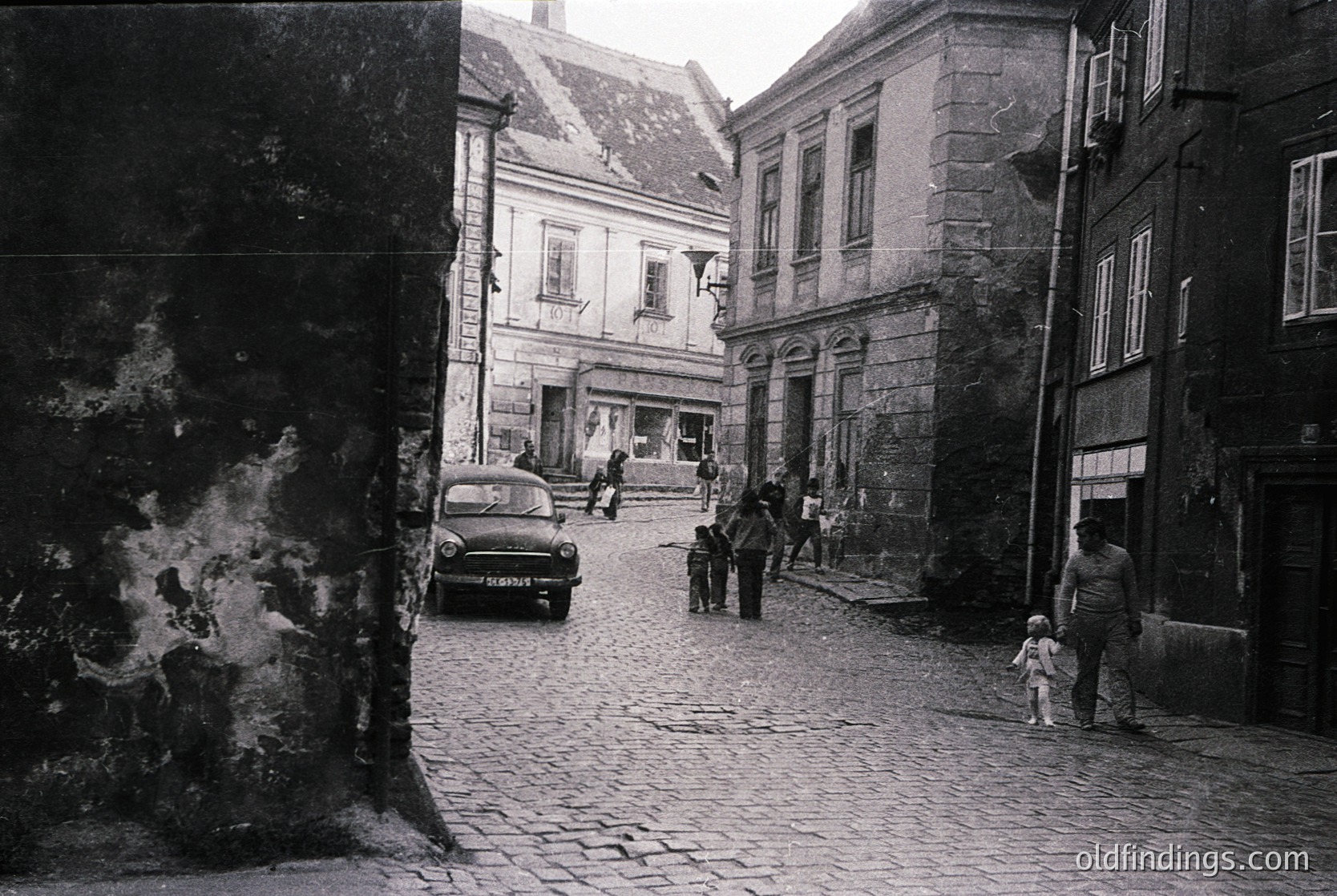 Mid-20th century European street scene with cobblestone pavement, vintage car (likely or ), and stone buildings. People dressed in 1960s-70s attire. Urban decay visible on left wall.