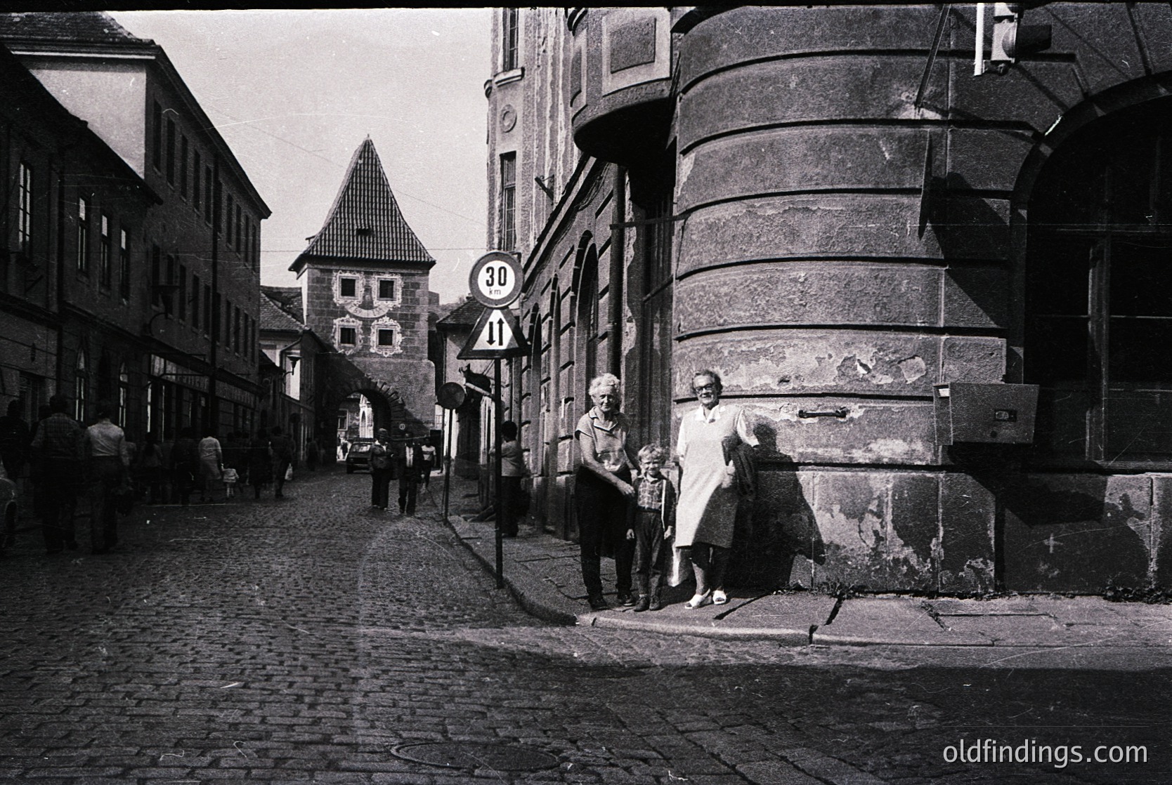 Mid-20th century European street scene with cobblestone pavement, featuring a woman in a long dress holding a child’s hand. Distinctive medieval-style tower and rounded corner building with visible wear. Speed limit sign (30) and vintage signage visible. Likely