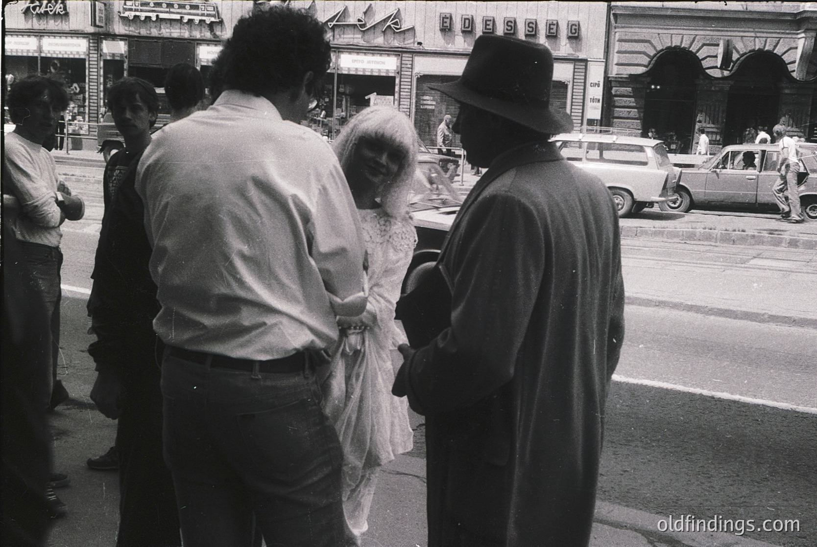 Black-and-white street scene featuring three central figures: a woman in a long, sheer wedding gown adjusting her veil, flanked by a man in a light shirt and another in a dark coat & hat. Classic 1960s–70s urban attire and vintage cars in background suggest a European city wedding. Candid, documentary-style capture.