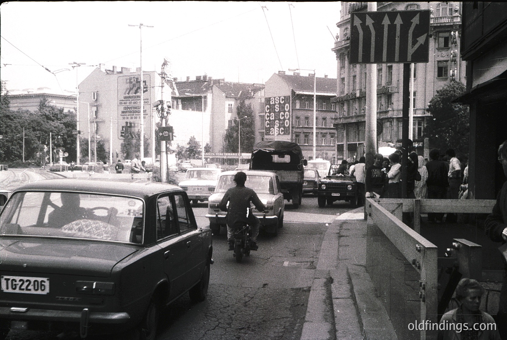 Urban street scene with mid-20th century vehicles, including a motorcycle and vintage cars. Pedestrians cross near a pedestrian bridge with "CSCO" signage. Soviet-style architecture and propaganda posters dominate the background. Likely Eastern Bloc, 1960s-1980s.