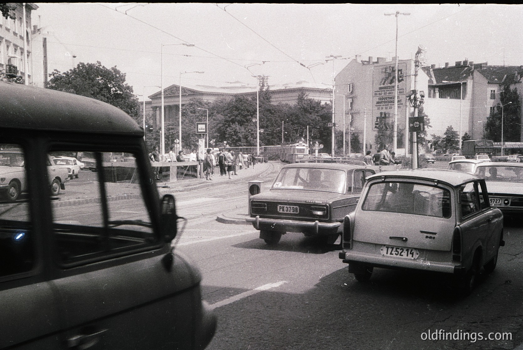 Vintage urban street scene with mid-20th century Soviet-era cars (Lada/ZAZ) and a bus, likely Eastern Bloc. Pedestrians cross at a traffic light near a large, classical-style building with columns. Posters on poles suggest state propaganda or public announcements.