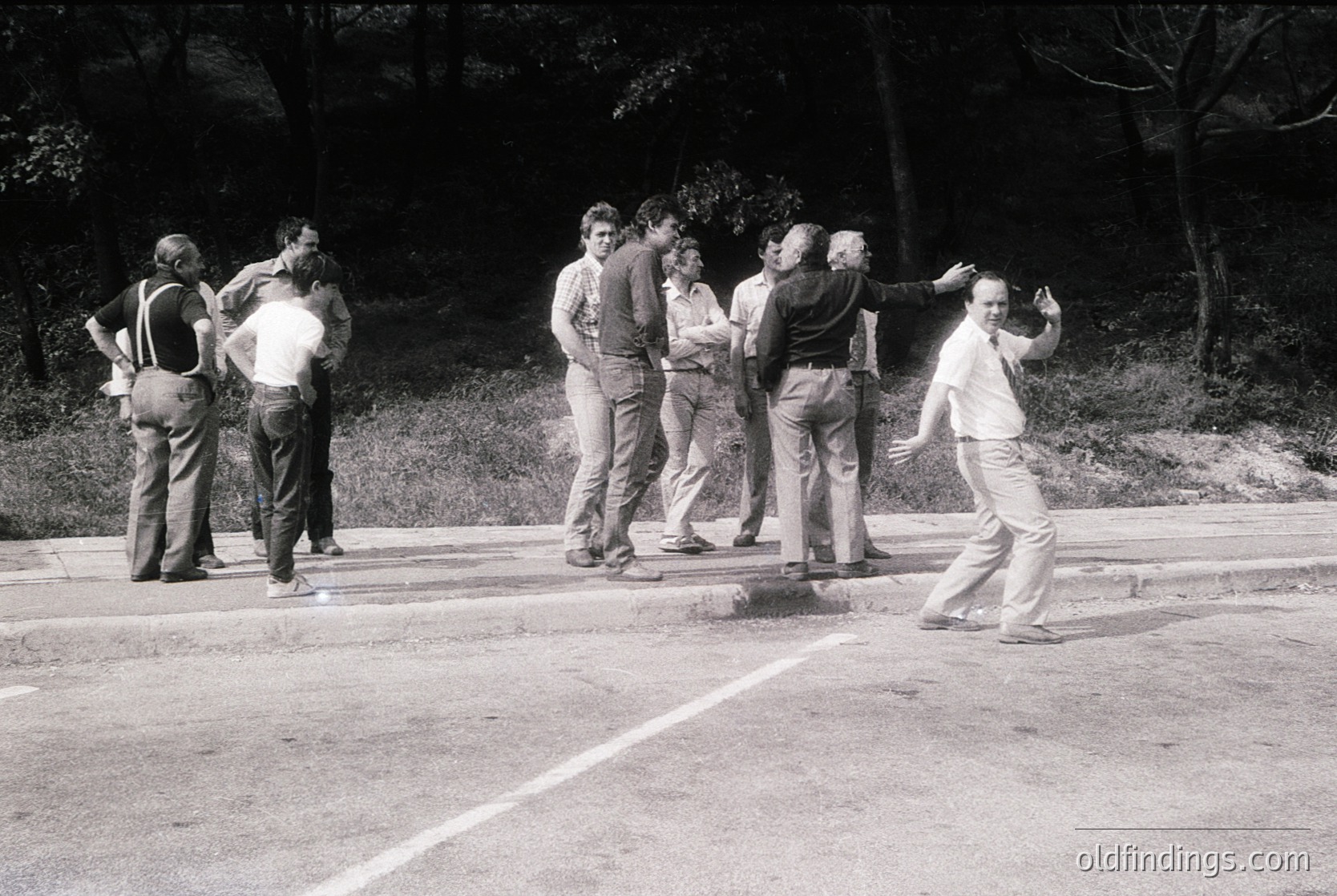 Group of men in casual 1970s attire playing frisbee on a paved area, likely a park or sports field. One man mid-throw, others watching or preparing to catch. Trees and greenery in background suggest an outdoor setting. Black-and-white photo captures candid, informal recreation.
