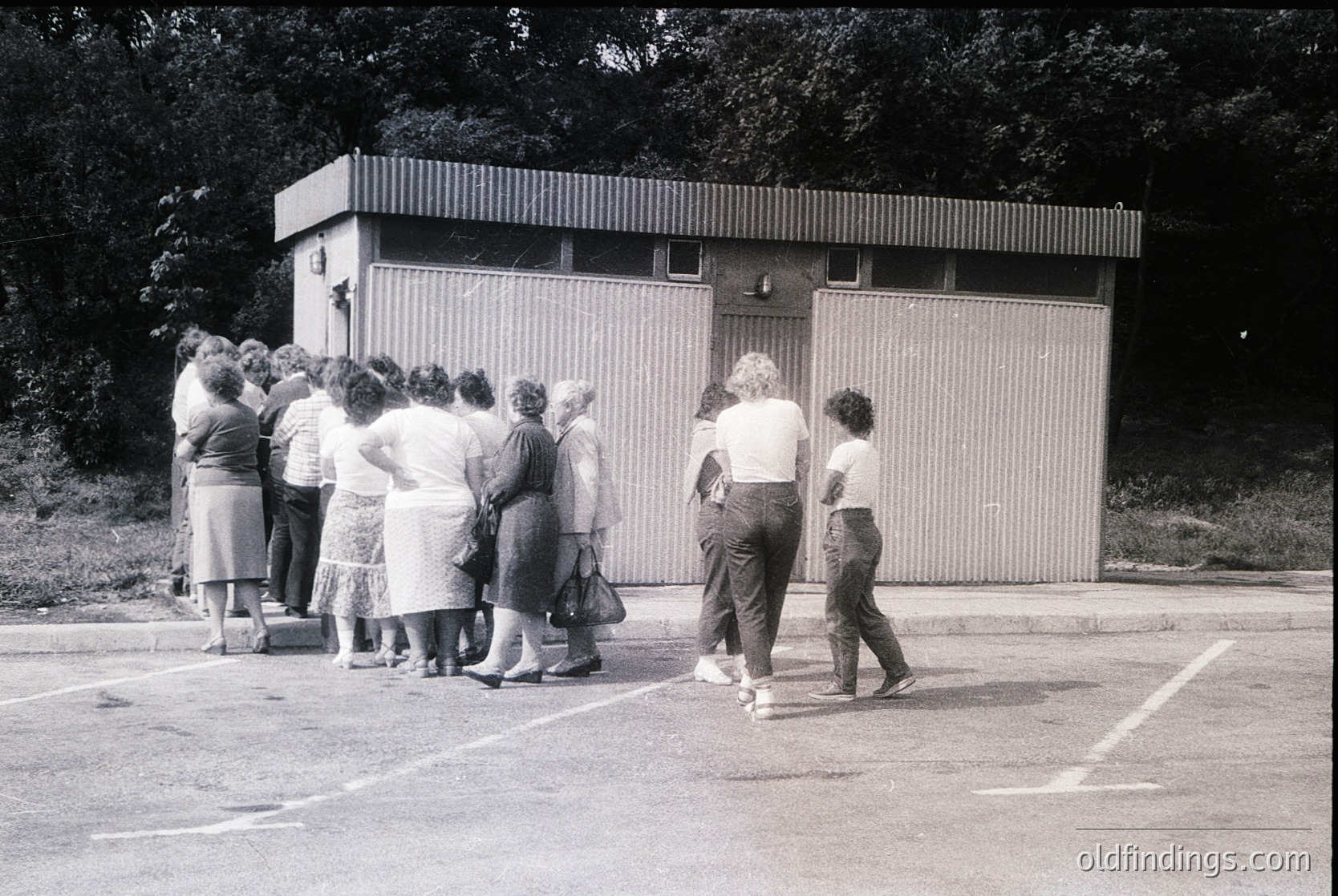 Mid-20th century group photo outside a utilitarian concrete block changing room. Women in 1960s-70s fashion—pleated skirts, blouses, and knee-high socks—wait in line. Lush greenery and trees frame the scene, suggesting a park or recreational area.