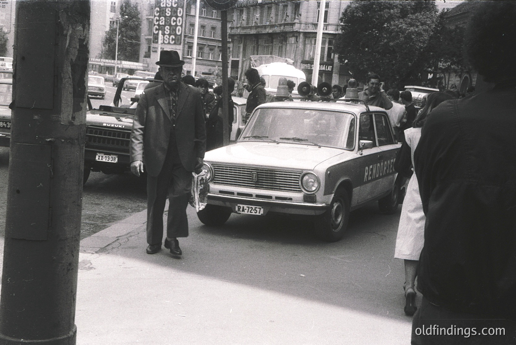 Vintage black-and-white street scene featuring a **1960s-era ambulance** (marked "ПЕРВАЙНА ПОМОЩ" in Cyrillic) parked on a busy urban road. A man in a **fedora and suit** walks beside it, while onlookers gather around. Soviet-era architecture and signage suggest Eastern Europe, likely **Bulgaria**. Crowds and parked cars indicate a bustling city atmosphere.