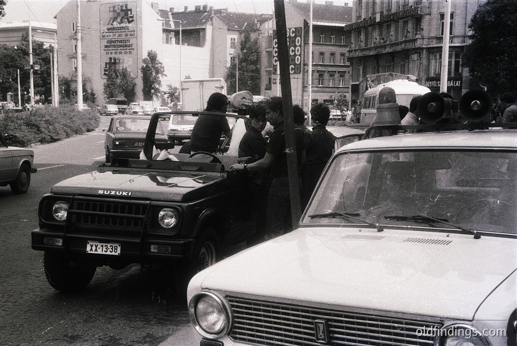 Black-and-white street scene featuring a Suzuki off-road vehicle with a group of people standing on its roof, likely during a public event or rally. A vintage white sedan (likely a Lada) and a white van are parked nearby. Soviet-era signage and urban architecture suggest Eastern Europe, mid-20th century (1970s–1980s).