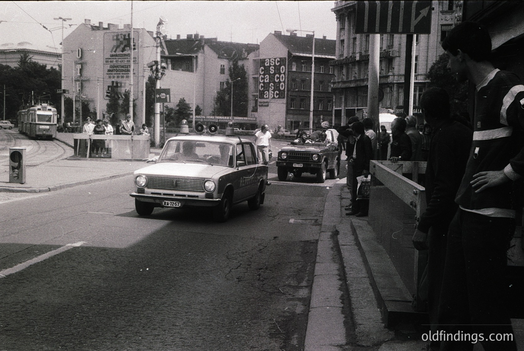 Vintage urban street scene featuring a 1970s-era sedan driving past a pedestrian bridge. Crowd on sidewalk, some wearing high-visibility vests. Prominent billboard reads "ASCO CASCO ASCO" on a multi-story building. Tram tracks and mid-century concrete architecture visible. Likely Eastern European city.