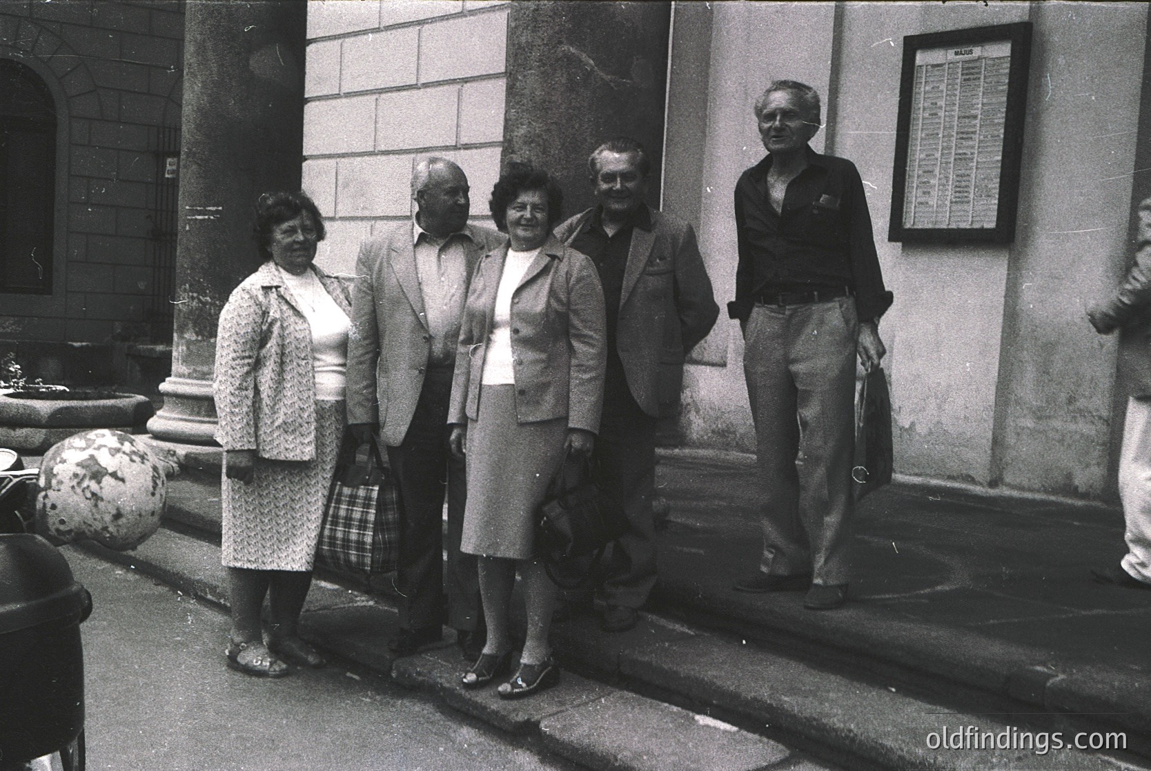 Black-and-white group portrait in front of a stone building, likely from the 1960s–1970s. Five adults pose formally, dressed in mid-century attire: suits, blouses, and patterned dresses. A signboard with text (unreadable) hangs on the wall. Urban setting with architectural details like columns and steps.