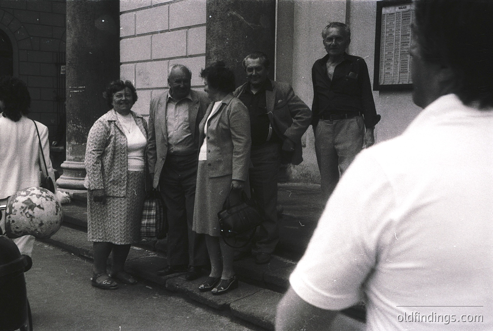 Group of six adults posing outside a building with classical architectural details, likely mid-20th century. Men wear suits/vests; women in patterned dresses and aprons. One woman holds a floral-patterned bag. Black-and-white photo suggests 1950s–1960s era.