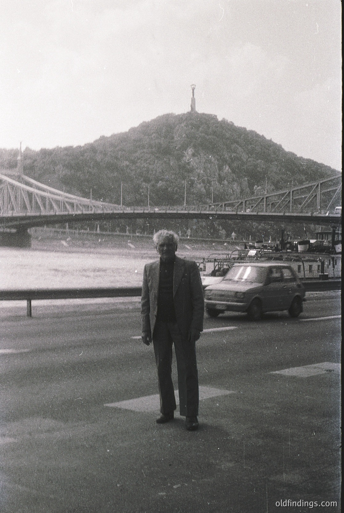 Mid-20th century black-and-white photo: Man in 1960s-era suit stands on a paved plaza beside a bridge, with a prominent hilltop statue and parked cars in background. Likely Lisbon’s 25 de Abril Bridge and Cristo Rei statue.