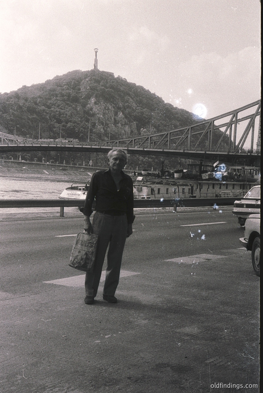 Mid-20th century black-and-white photo of a man standing near a bridge with a statue atop a hill in the background. He holds a patterned bag and wears a suit, suggesting a formal or transitional era. Industrial bridge and parked vehicles indicate urban transport infrastructure. Likely Eastern Europe, 1950s-1960s.