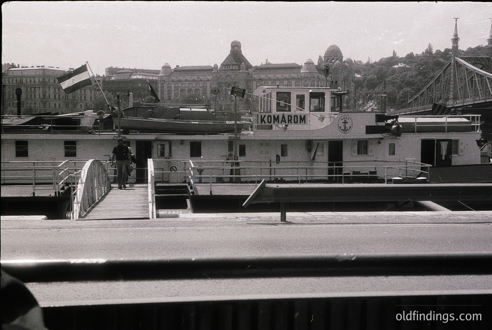 Black-and-white ferry docked at Budapest’s Danube River with "Ikomarom" branding, mid-20th century. Historic cityscape of domes and bridges in background, including Parliament. Single passenger on metal gangway, flag atop vessel.