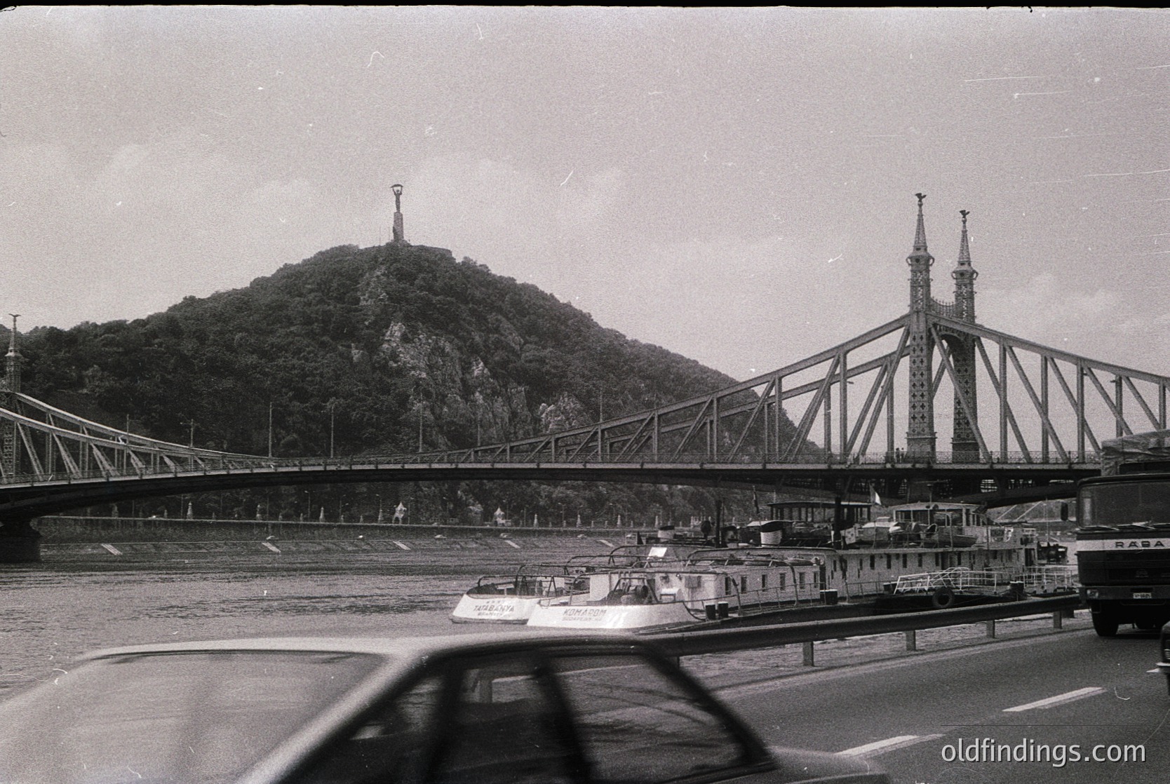 Black-and-white shot of the Liberty Bridge (Szabadság híd) in Budapest, Hungary, spanning the Danube River. The bridge’s iconic truss design and spires frame a hilltop statue. Mid-20th century (1950s–1960s) urban transport scene with vintage cars, riverboats, and pedestrians.