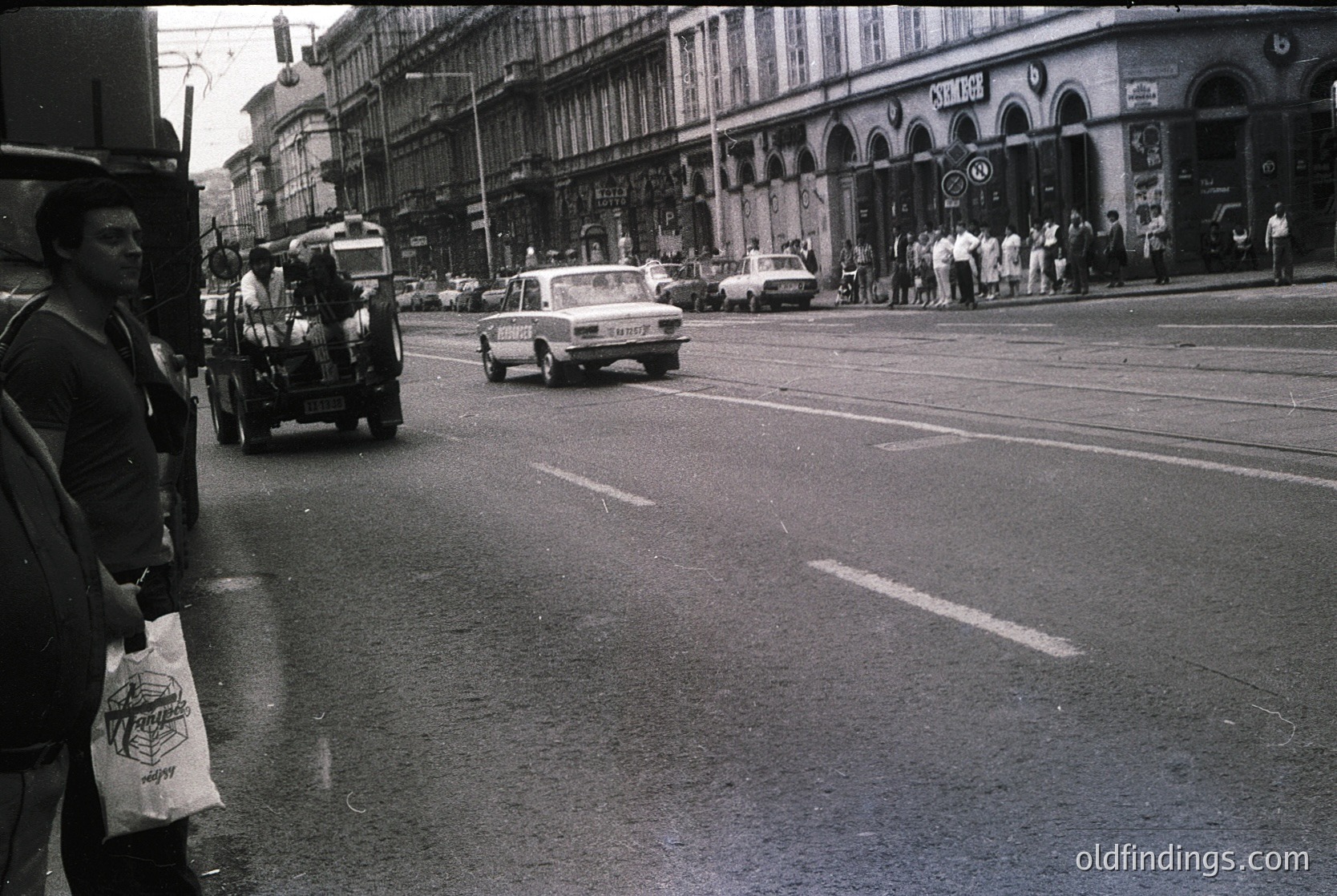 Mid-20th century urban street scene with vintage vehicles, including a horse-drawn cart and classic cars. Pedestrians line the sidewalk near a row of European-style buildings with arched storefronts. Tram tracks run through the road, indicating public transit infrastructure. Likely Eastern Bloc or Western European city, 1950s–1970s.