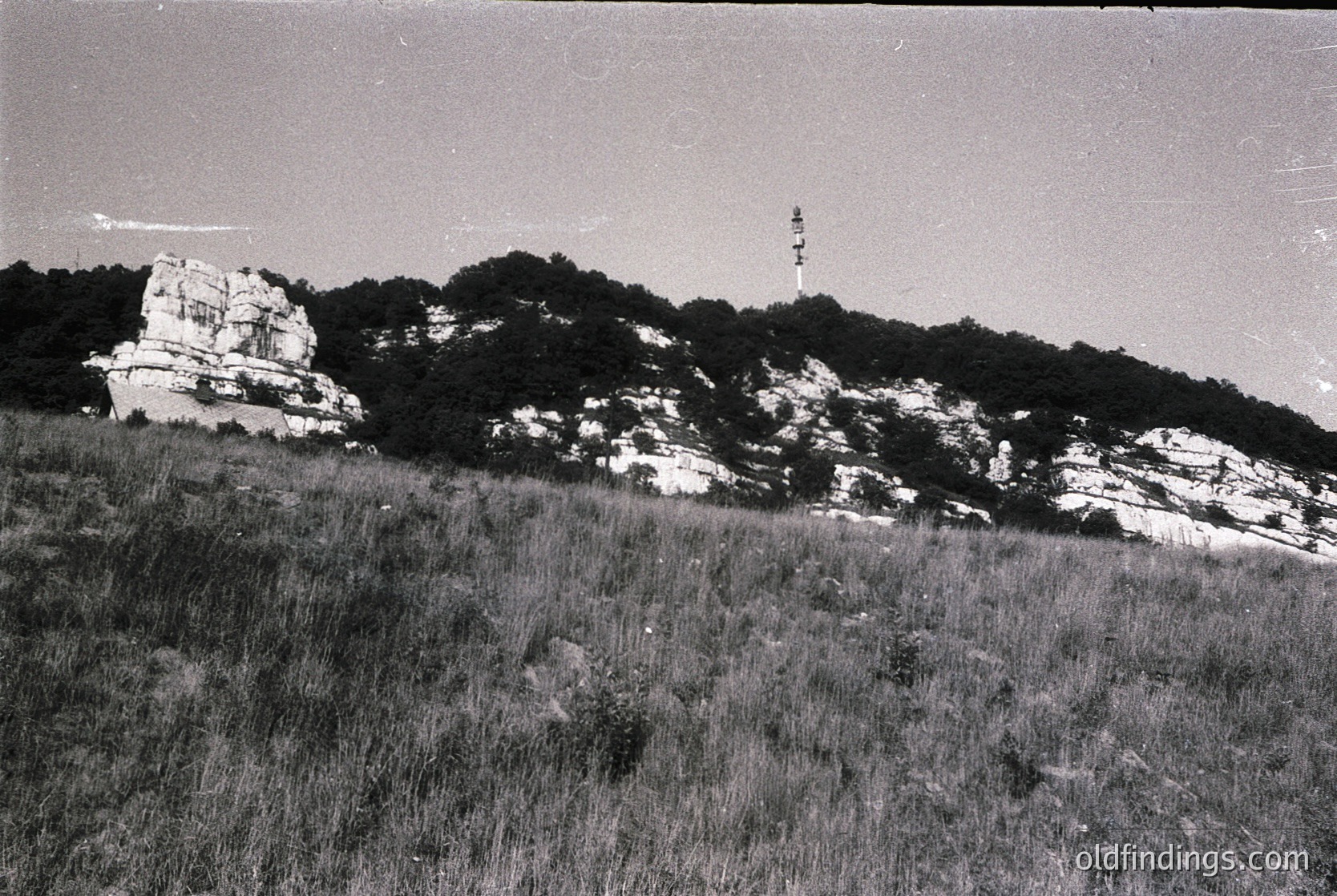 Vintage black-and-white coastal landscape featuring rugged limestone cliffs and a tall lighthouse on a hilltop. Dense vegetation covers the lower slopes, with sparse grassy areas in the foreground. The lighthouse structure appears industrial, likely mid-20th century.