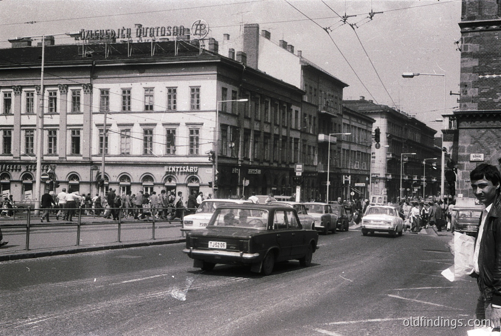 Vintage urban street scene featuring early 20th-century European architecture with a prominent three-story building displaying Cyrillic signage ("Друмелица Дуросач"). Tram lines and vintage cars (Lada, Škoda) navigate a busy intersection. Pedestrians in 1960s-70s attire cross sidewalks lined with shops and cafés. Overcast sky and urban infrastructure suggest a Soviet-era city.