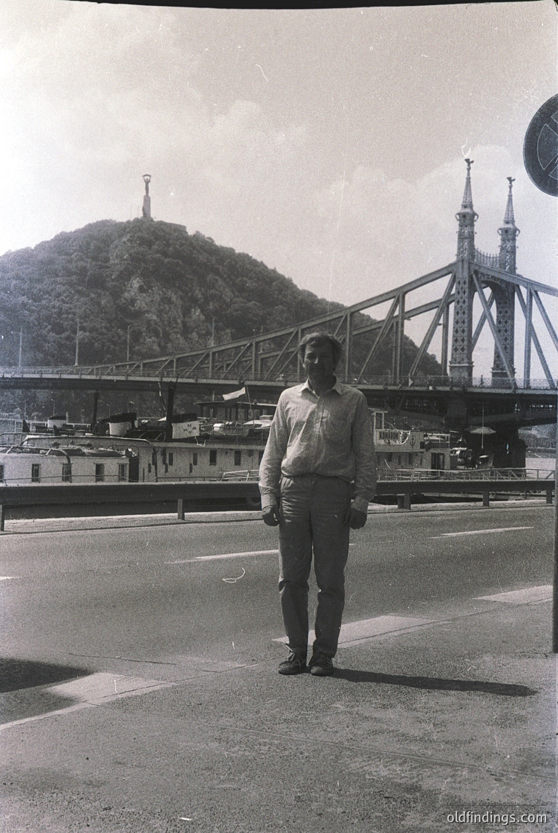 Man in 1960s-style suit stands beside the **Vitosha Bridge (Sofia, Bulgaria)**, a Soviet-era steel arch structure. Industrial port and hilltop monument visible in background. Black-and-white, mid-century urban scene.