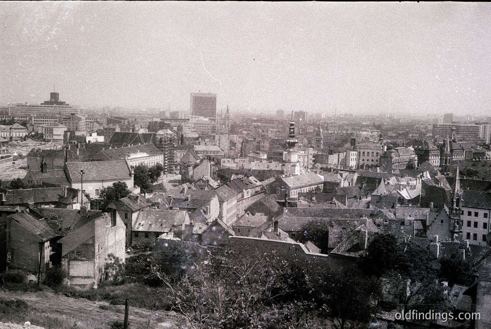 Black-and-white aerial view of a European cityscape, likely early-to-mid 20th century. Dense cluster of low-rise buildings with pitched roofs, mixed with taller structures and church spires. Prominent central square or plaza with a large, symmetrical building. Industrial-era architecture with visible chimneys and utility lines. Fog or haze obscures distant skyline.