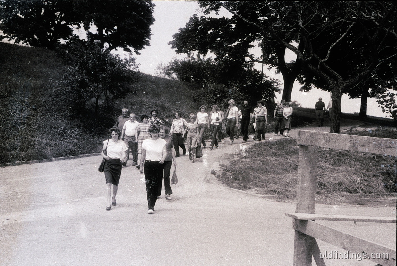 Group of people walking along a tree-lined path, likely a public park or promenade. Mid-20th century clothing (1950s–1960s) suggests casual, leisurely activity. Concrete steps and wooden railings indicate urban park design.