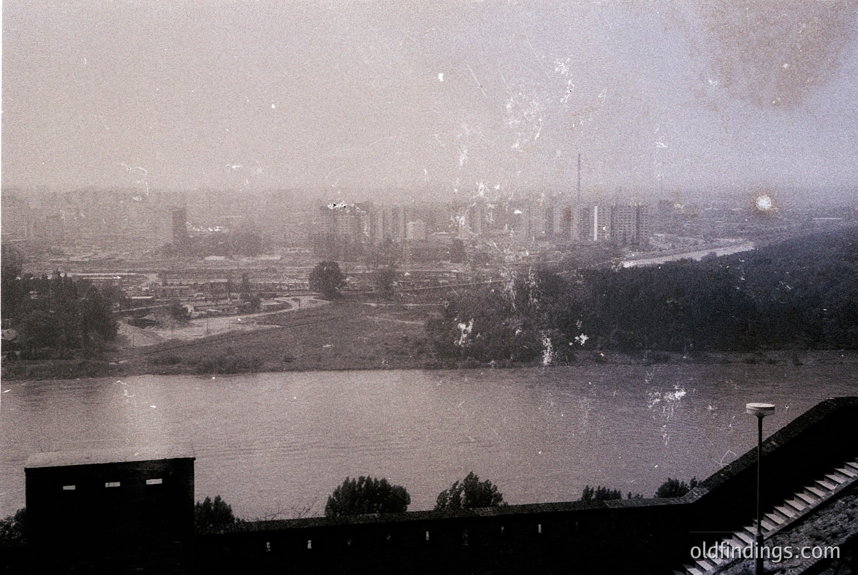 Vintage sepia-toned aerial view of an industrial riverside cityscape. Prominent brick factory buildings line the riverbank, with smokestacks and cooling towers visible. Dense urban sprawl extends beyond, featuring grid-patterned streets and scattered greenery. Likely Eastern European industrial zone, mid-20th century.