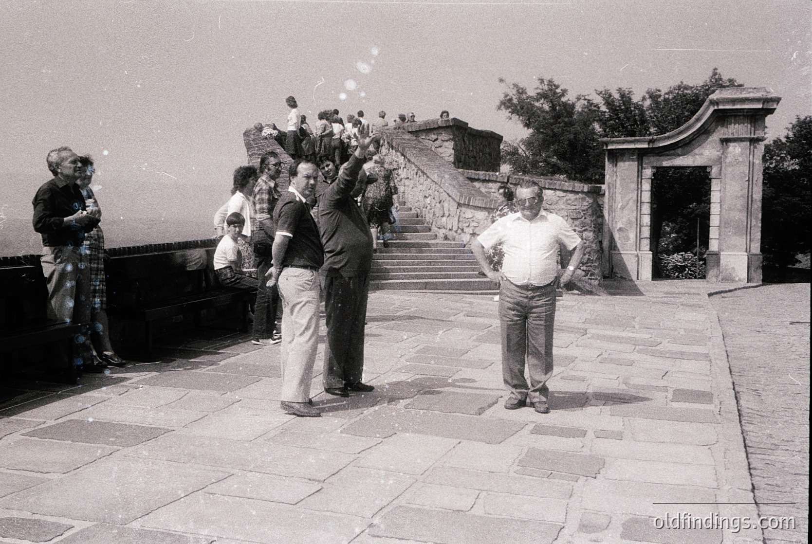 Black-and-white snapshot of a mid-20th-century gathering at a grand stone staircase with classical arches. A man in a white shirt and light trousers poses confidently, while others—including a child and adults in casual 1960s-70s attire—stand or sit on the steps. One man throws confetti, suggesting a celebratory or festive occasion. Lush greenery and distant cityscape visible in background.