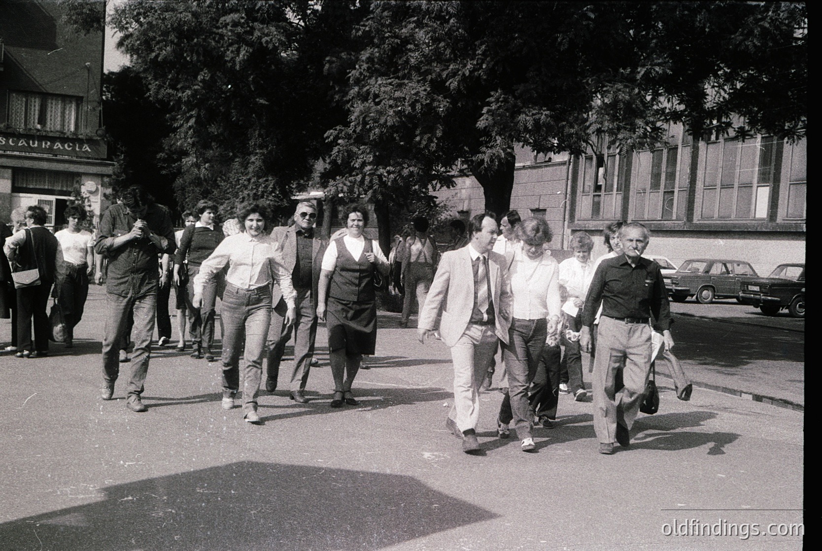 1970s street scene in a European city, likely Spain. Group of casually dressed pedestrians—men in suits, women in blouses and skirts—walking near a "GLAU PACIA" storefront. Classic mid-century architecture with parked vintage cars. Urban lifestyle documentation.