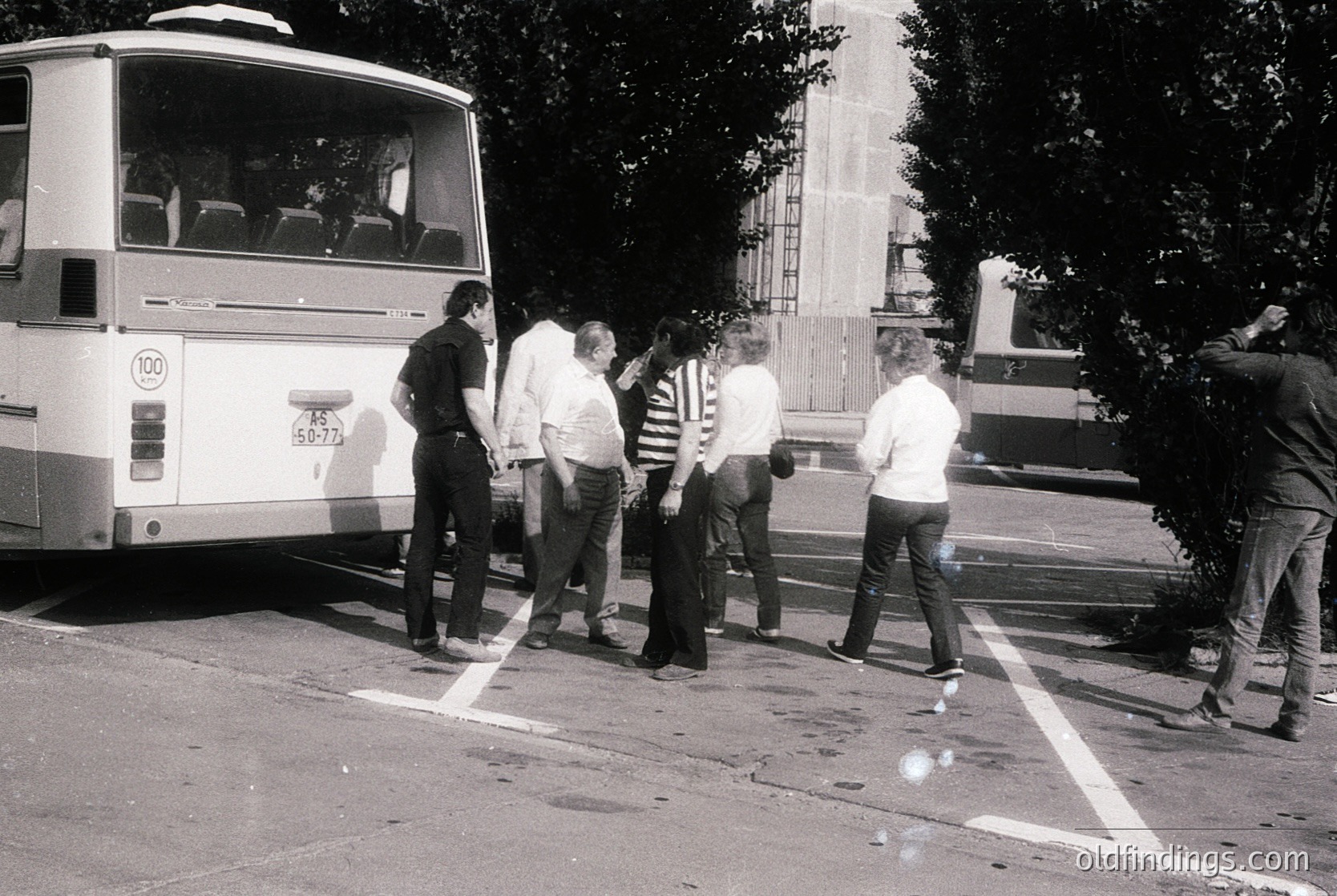 Group of five men in 1970s-era streetwear (striped shirts, bell-bottoms) standing near a white bus marked "100" in a parking lot. Urban setting with visible road markings, trees, and a monument in background. Likely Eastern Bloc-era public transport scene.