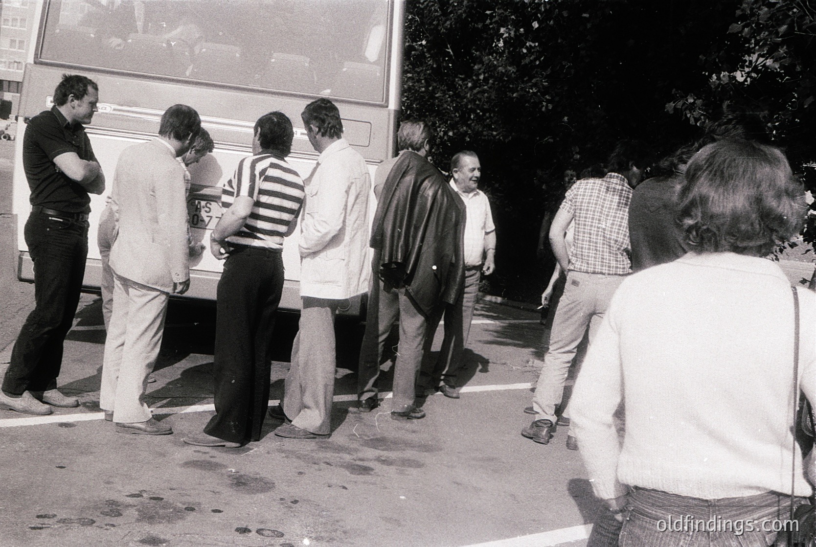 Black-and-white street scene: group of 7 adults in 1970s-era clothing (striped shirts, button-downs) standing in line at a bus stop under a tree. Urban setting with visible pavement and parked vehicles.