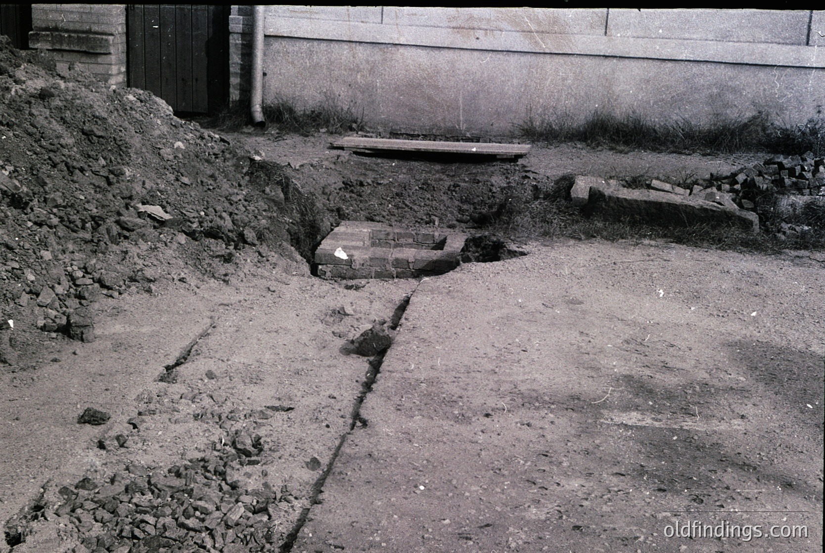 Black-and-white urban excavation site showing exposed brickwork and concrete remnants. Pile of rubble and dirt alongside a partially dug trench, likely for infrastructure repair or construction. Mid-century architectural context suggested by materials and layout.