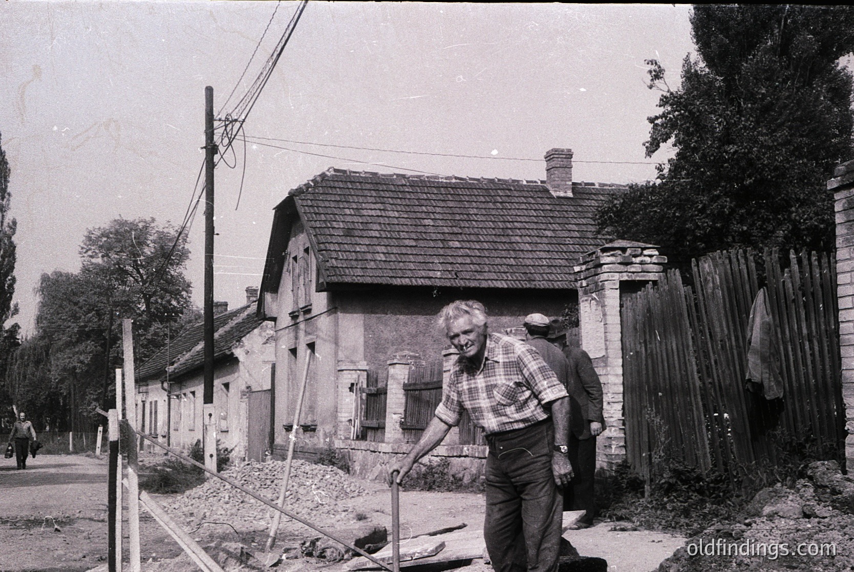Vintage black-and-white photo of a man in a plaid shirt and overalls holding a shovel, standing near a partially demolished brick house with exposed wooden beams. Overgrown vegetation and debris surround the site, indicating post-war or transitional-era reconstruction. Urban decay and rural architecture blend in this mid-20th-century setting.