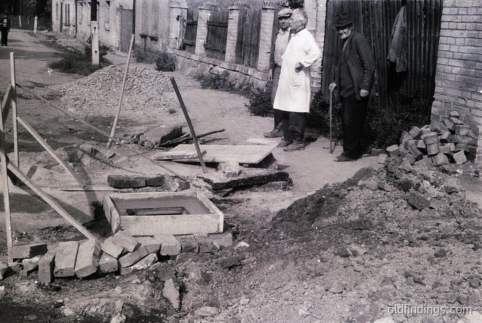 Mid-20th century street scene showing two men in formal attire observing a partially demolished brick wall and exposed foundation. Wooden scaffolding and rubble indicate construction or demolition work. Urban setting with adjacent buildings and unpaved ground.