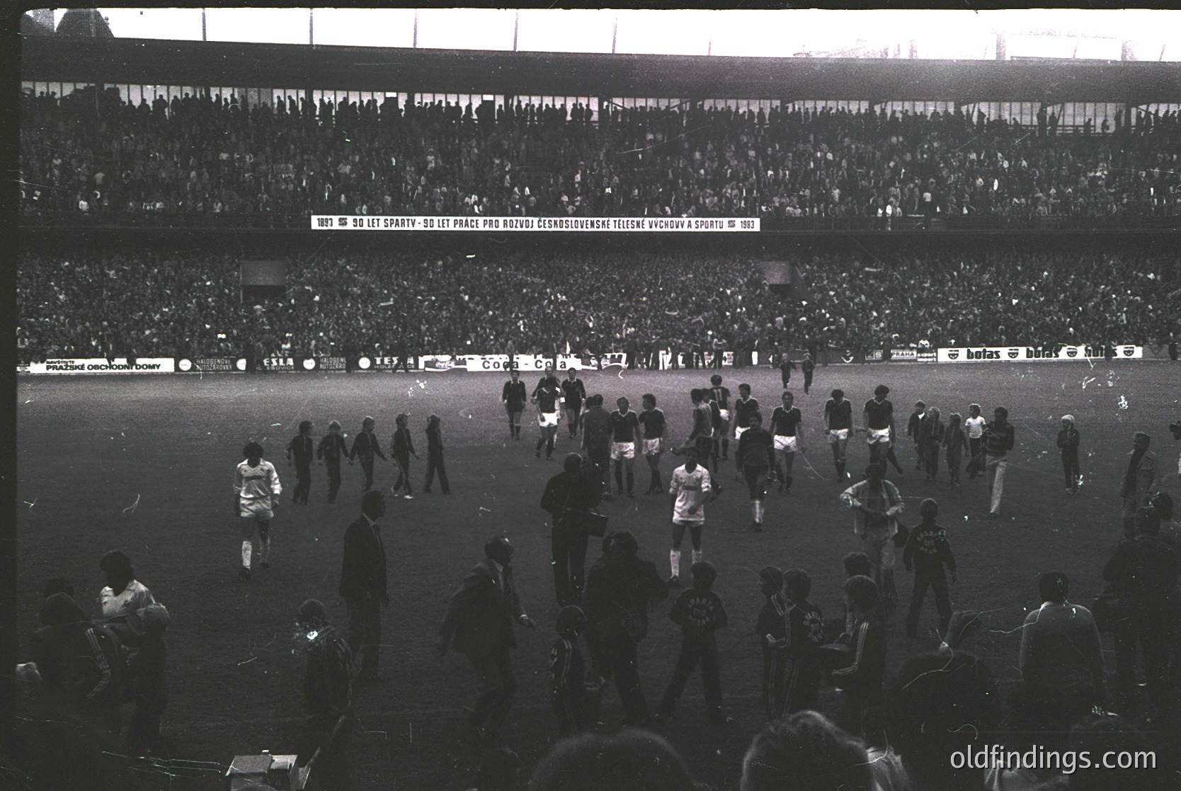 Black-and-white stadium scene capturing post-match chaos, 1960s-70s. Crowd surges onto field; players and spectators mix amid empty goalposts. Banner in Cyrillic suggests Eastern European location. Iconic for sports history research.